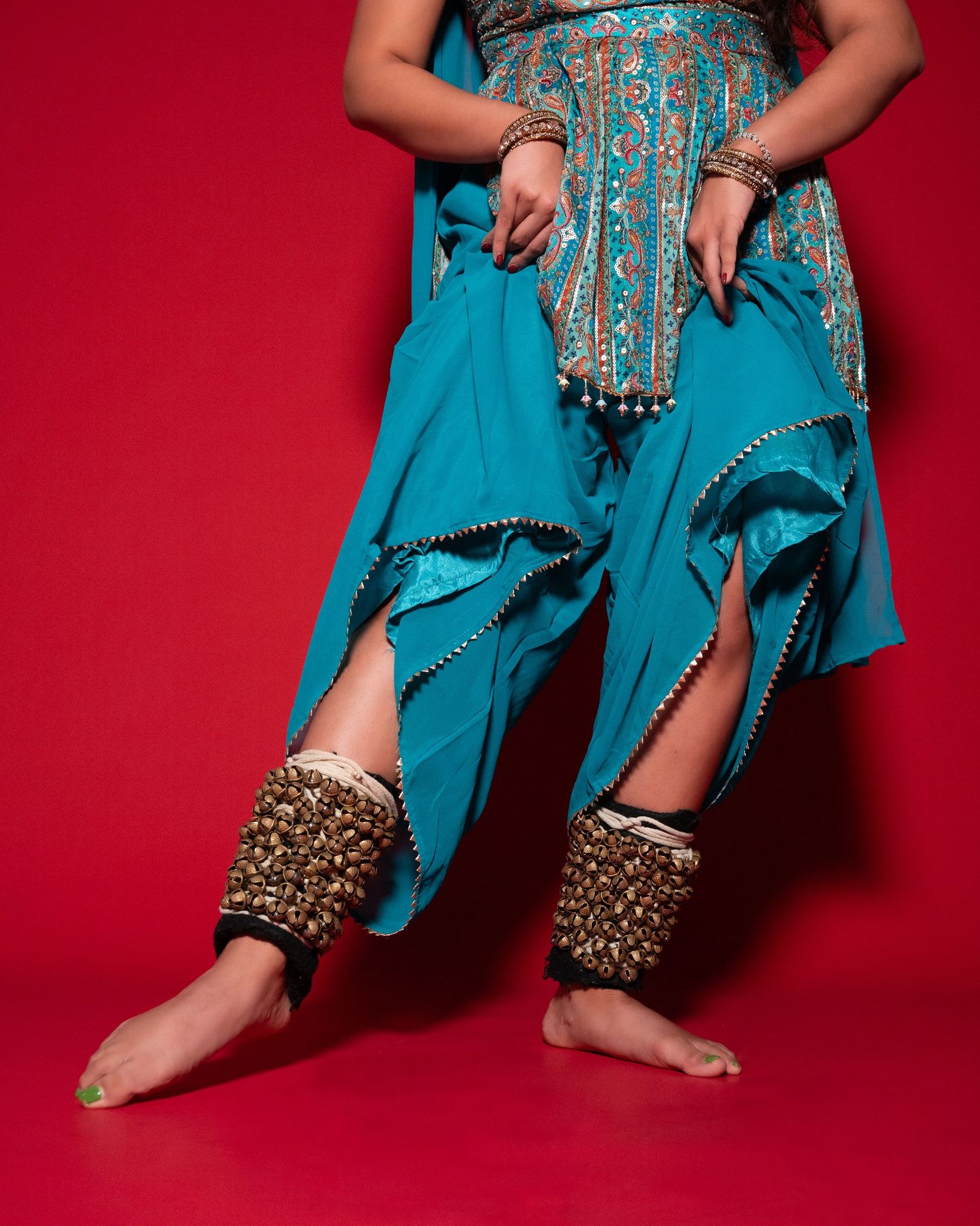 Dancer in teal harem pants with gold embroidered anklets poses against a red background.