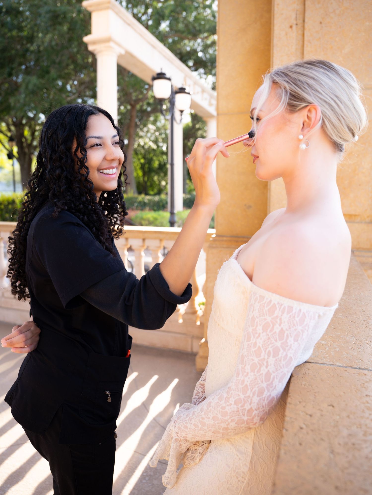 A makeup artist in black applies makeup to a blonde bride in a white lace dress outdoors at an elegant venue.