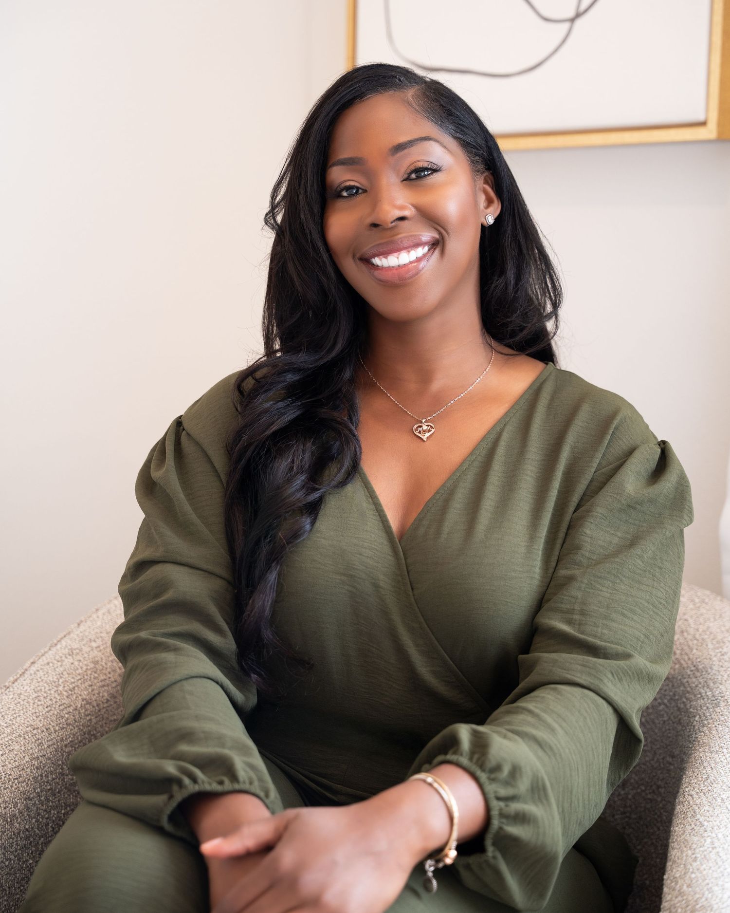 A smiling woman in an olive green wrap jumpsuit sits in a chair, wearing a gold necklace and bracelet.
