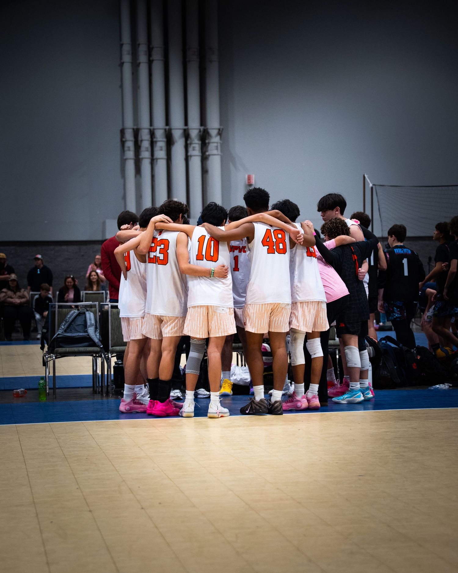 Youth volleyball team huddles together in white and orange uniforms on an indoor court during a game.
