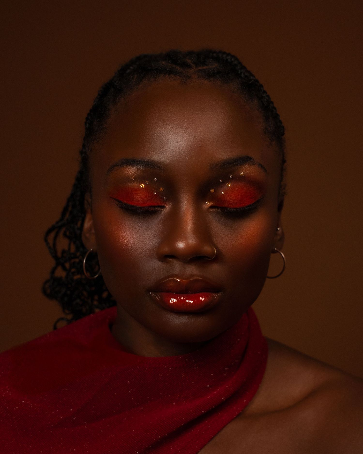 A Black woman with red glittery eye makeup and glossy lips poses in a red outfit against a brown background.