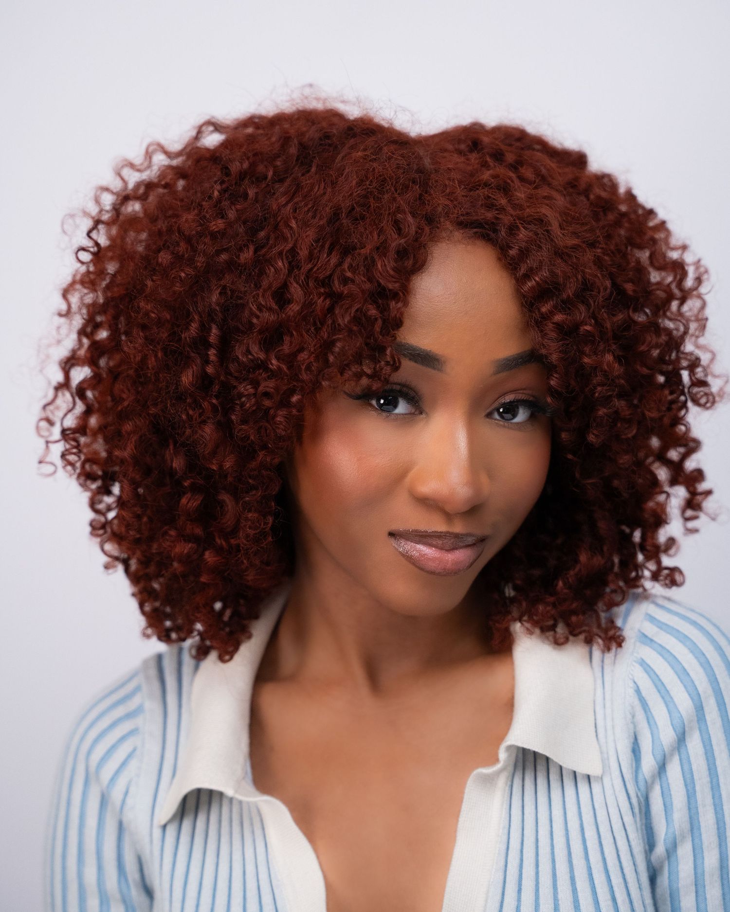 A young Black woman with voluminous auburn curly hair wearing a blue striped shirt poses against a white background.