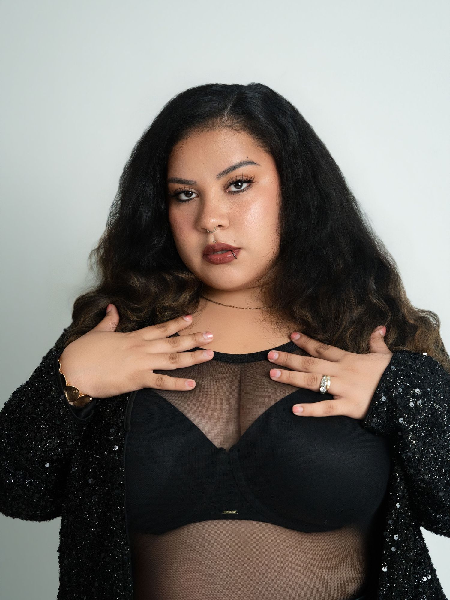 Woman in black bra and sequin jacket posing confidently against a light gray background.