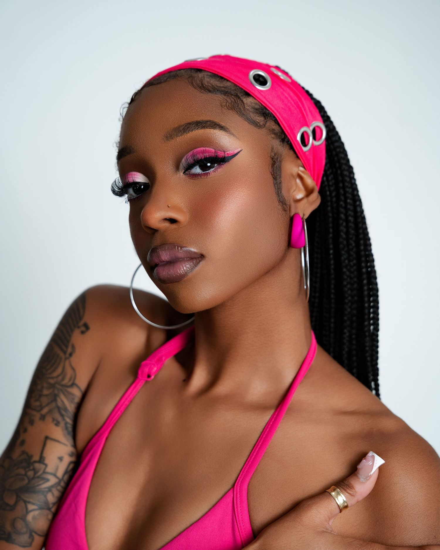 Woman in hot pink bikini top and headband with bold pink eye makeup, hoop earrings, and box braids poses confidently.