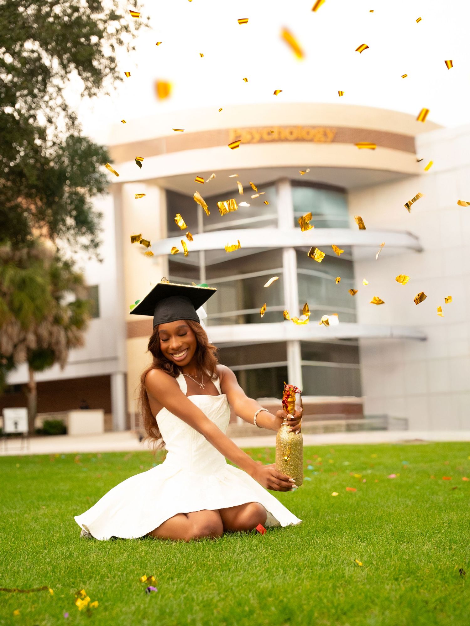Graduate in white dress and cap throws yellow flower petals while sitting on green grass during outdoor photo session.
