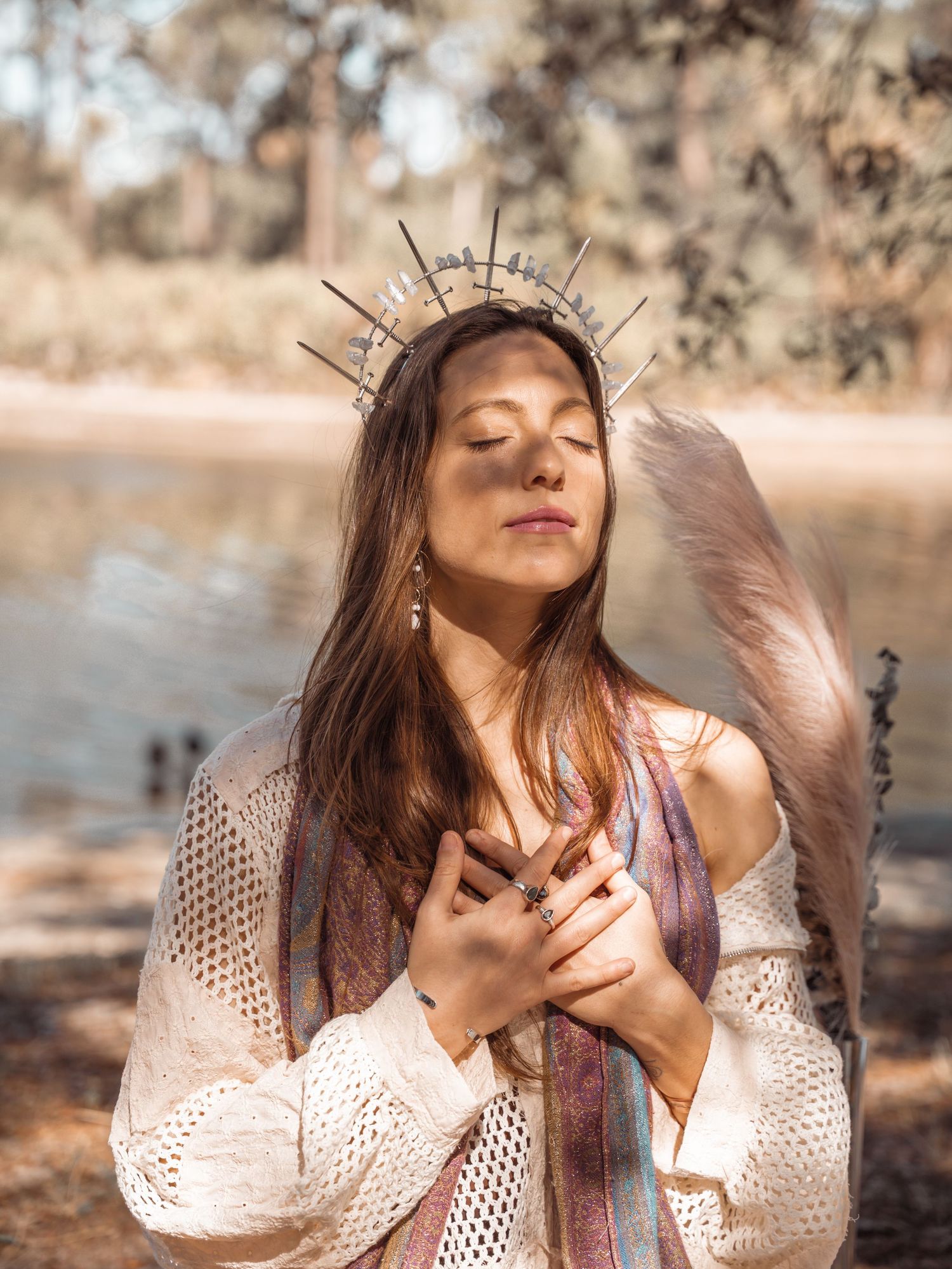 Woman wearing a crown and angel wings poses peacefully with eyes closed near a sunlit lake.