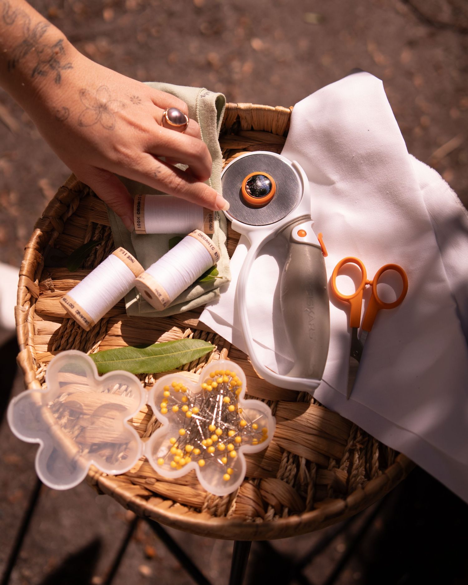 Tattooed hand arranging a wicker sewing basket with thread spools, orange scissors, pins, and an owl pincushion.