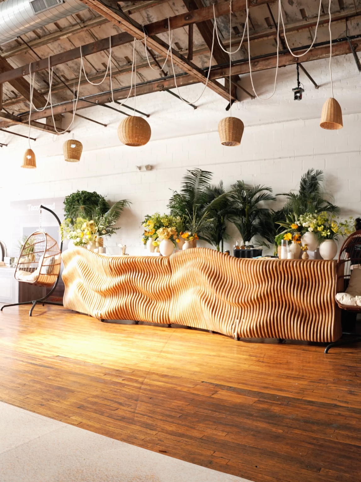 Modern event space with wavy wood bar, tropical plants, and hanging rattan pendant lights on hardwood floors.