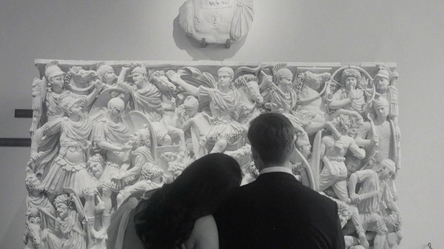 Couple embracing before an ornate Roman relief sculpture in a museum, black and white photo.