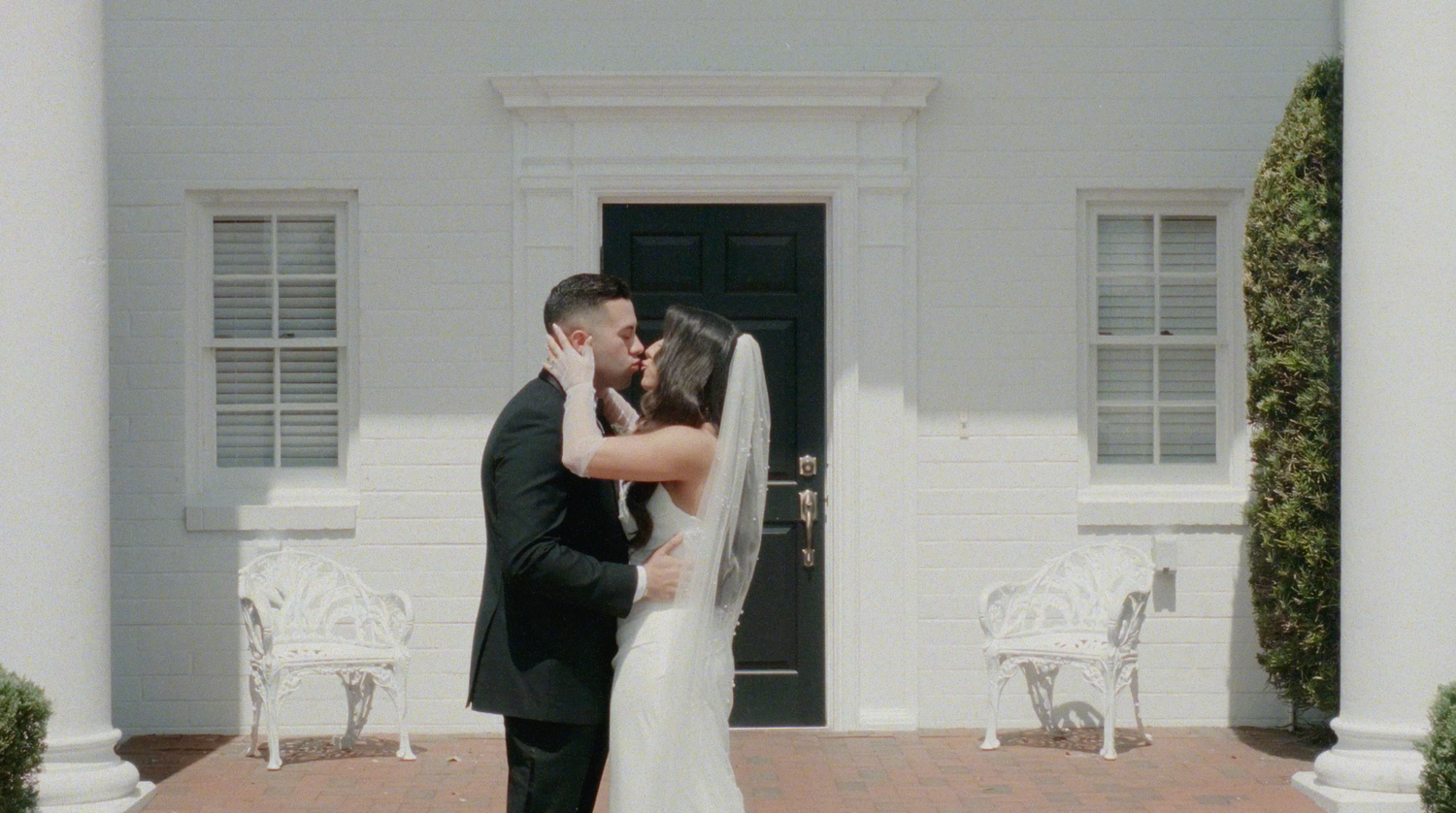 Bride and groom share a romantic kiss outside a white colonial venue with elegant white chairs.