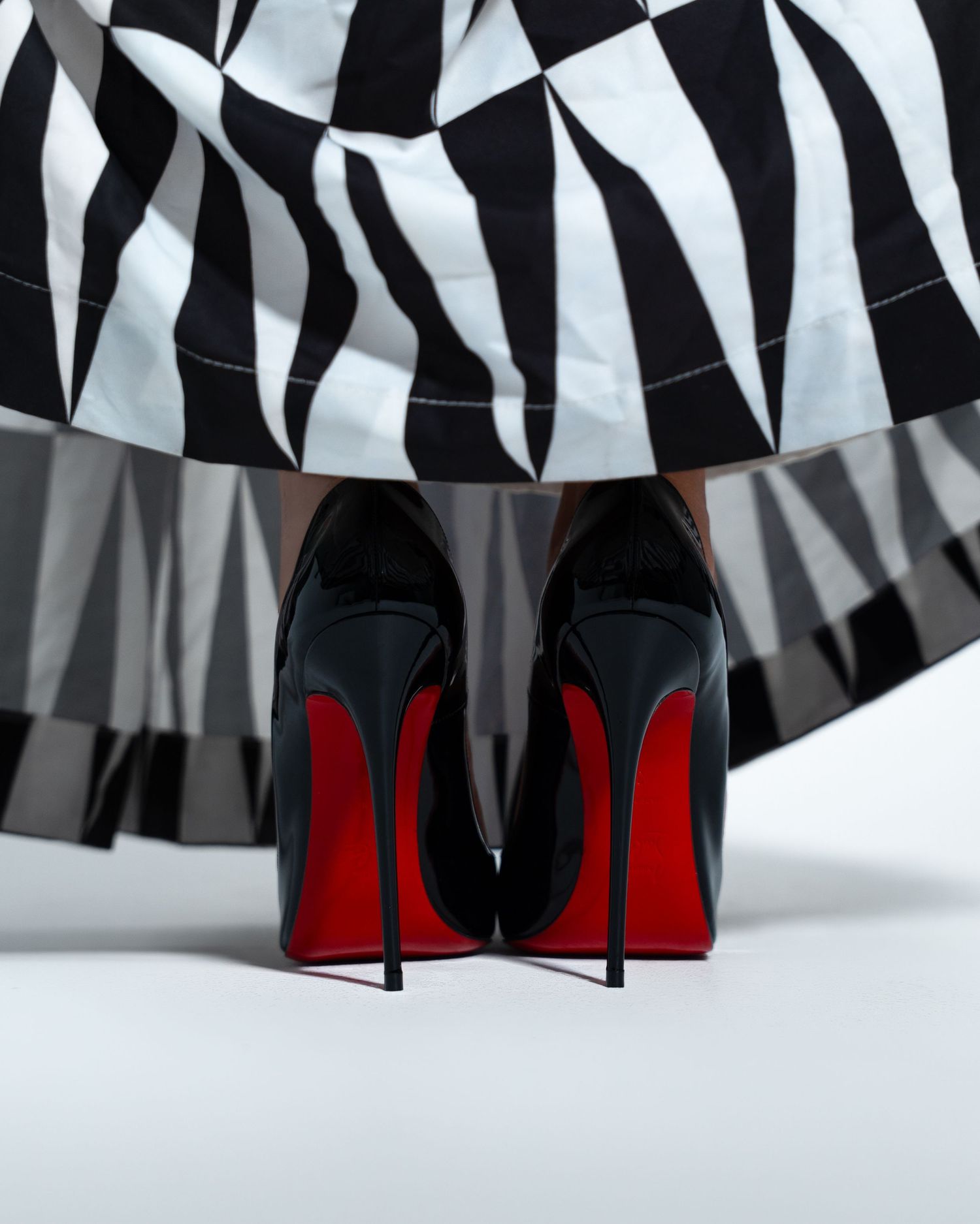 Black patent Christian Louboutin stiletto heels with iconic red soles beneath a black and white geometric skirt.