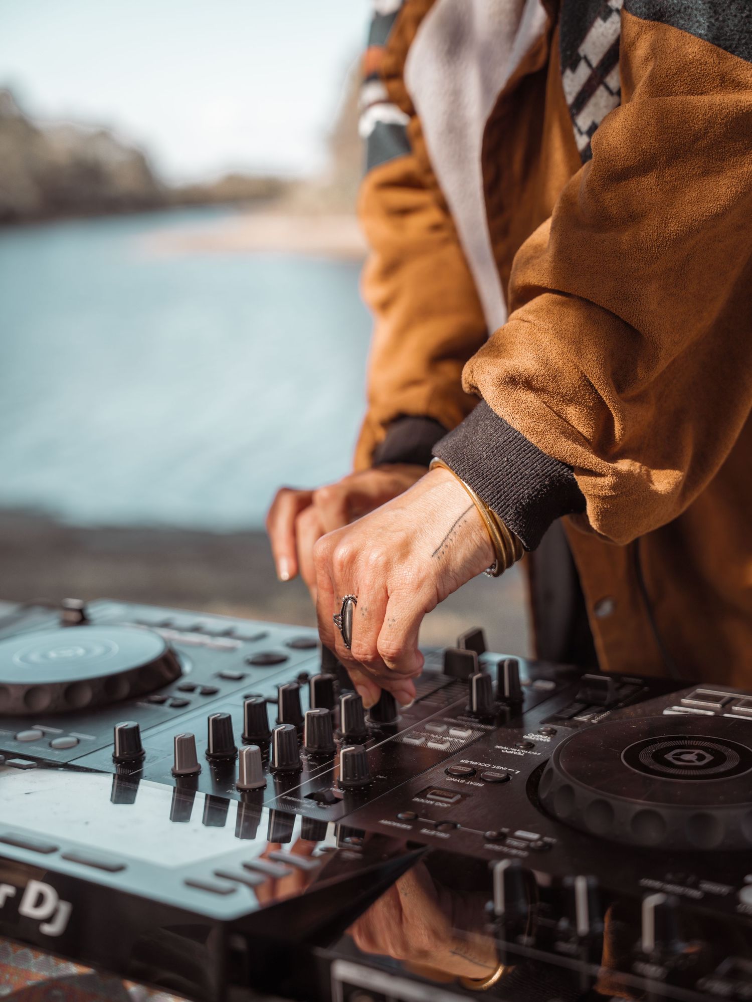 DJ mixing music outdoors on a Pioneer DJ controller near a waterfront, wearing a brown jacket.
