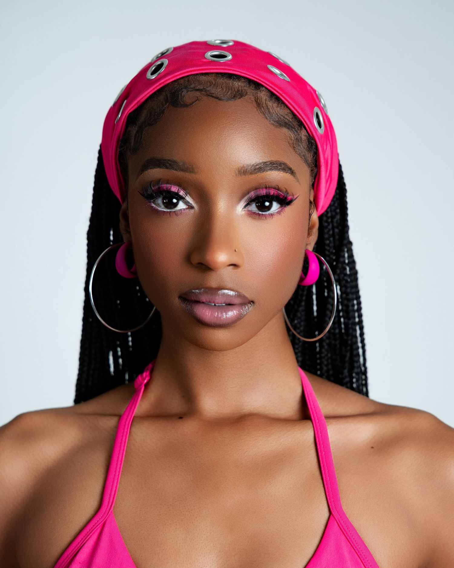 Young woman in hot pink halter top and headband with hoop earrings, wearing bold pink eye makeup and long braids.