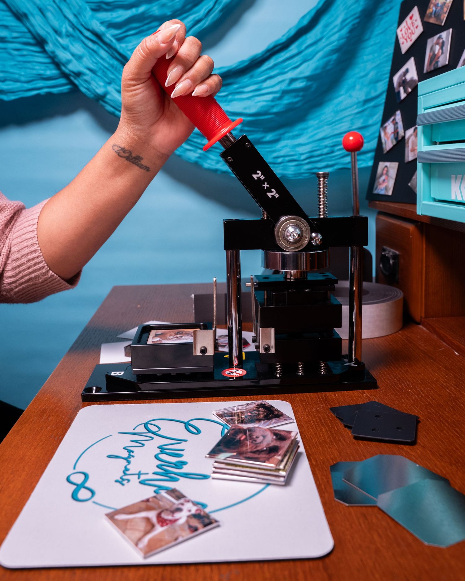 A woman uses a red-handled button maker press on a wooden desk with photo prints and a branded mousepad nearby.