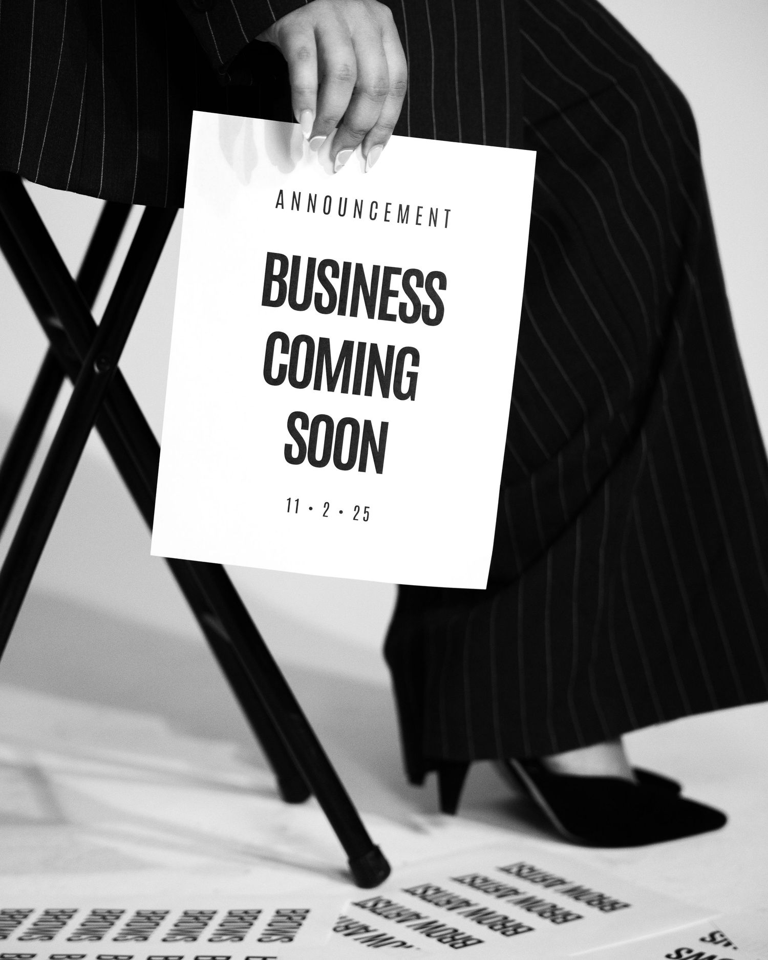Black and white photo of person in pinstripe suit holding Business Coming Soon announcement sign dated 11-2-25.