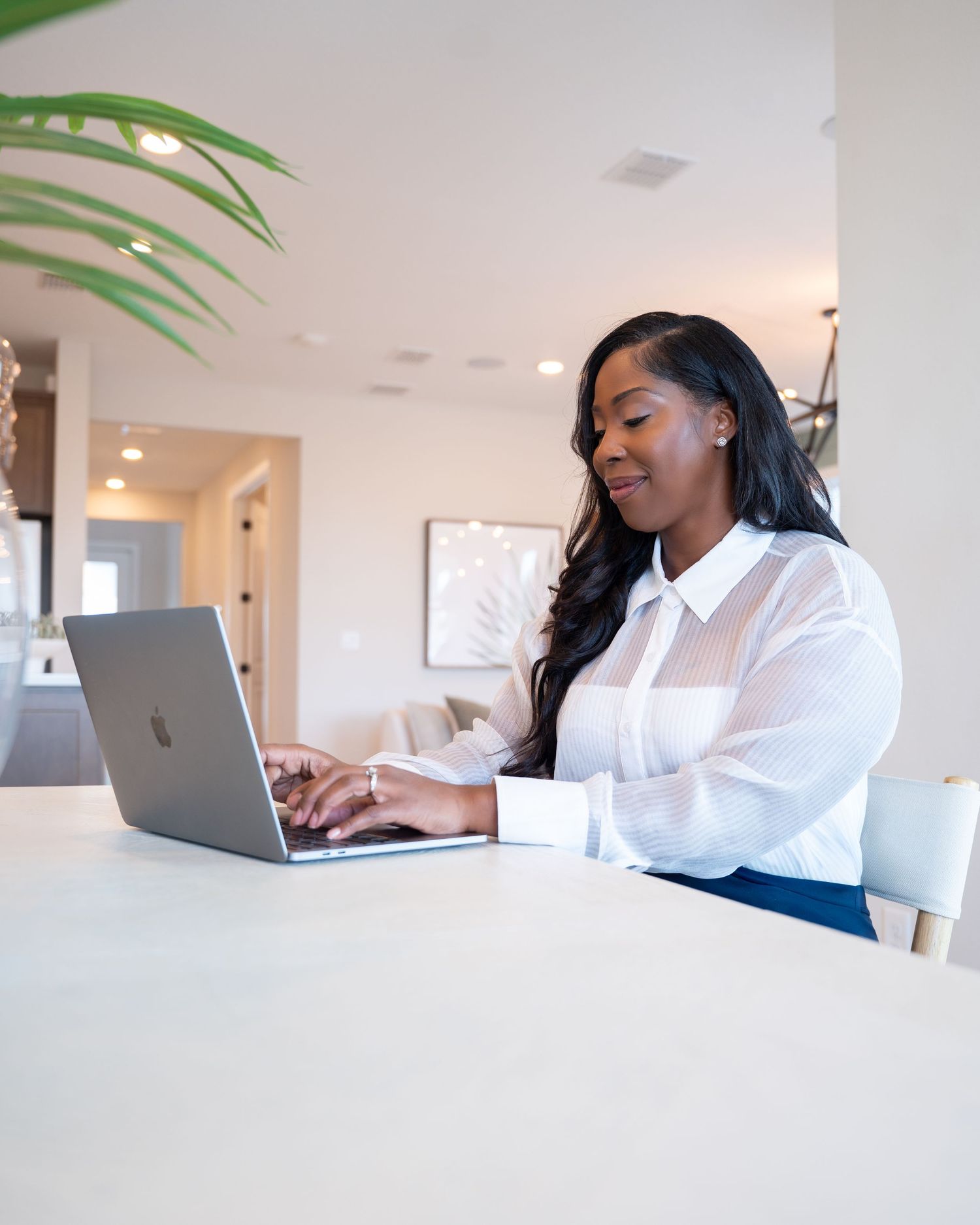 A young Black woman in a white blouse works on a MacBook laptop at a white table in a modern home.