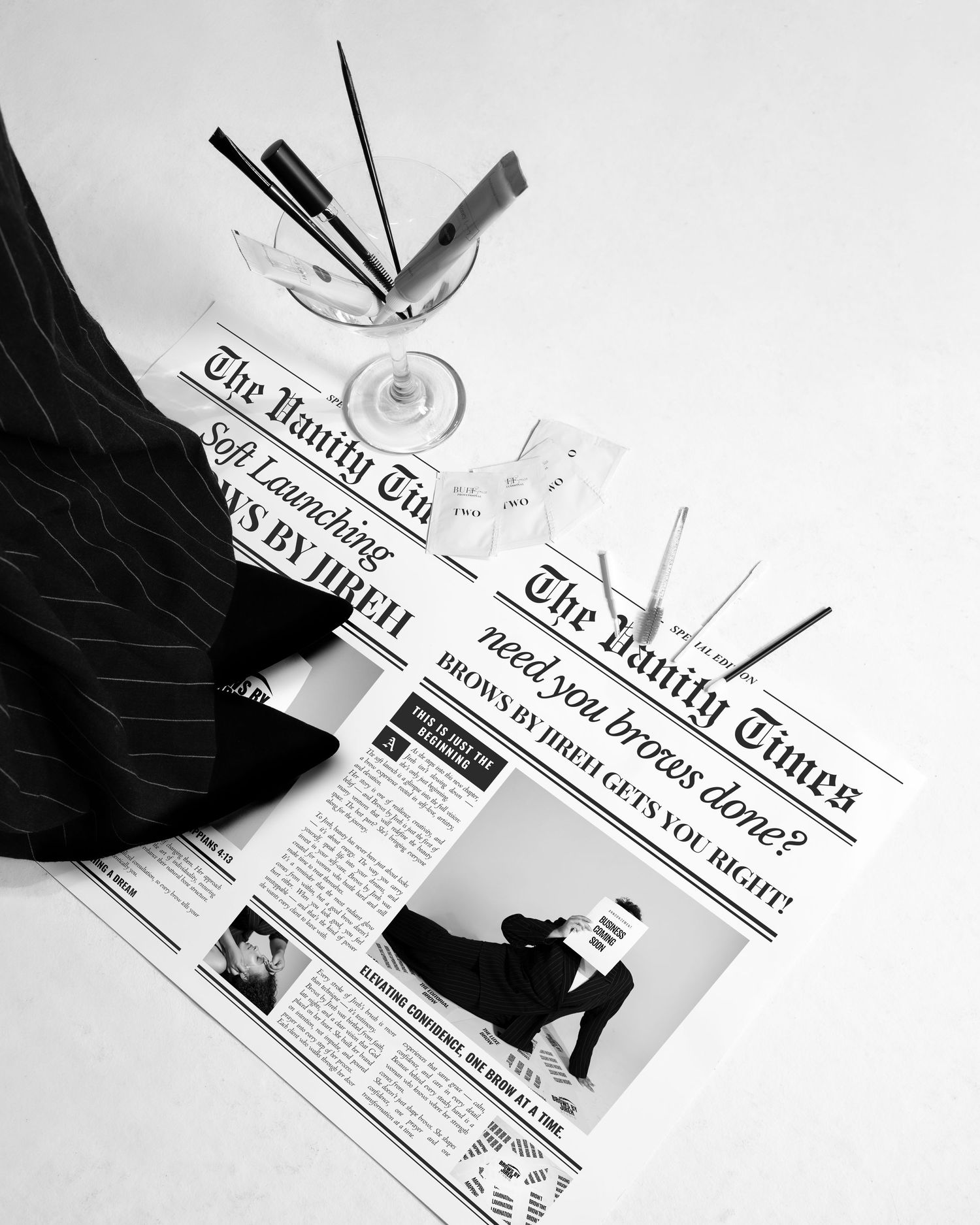 Black and white flat lay of The Vanity Times newspaper with makeup brushes in a glass on white surface.