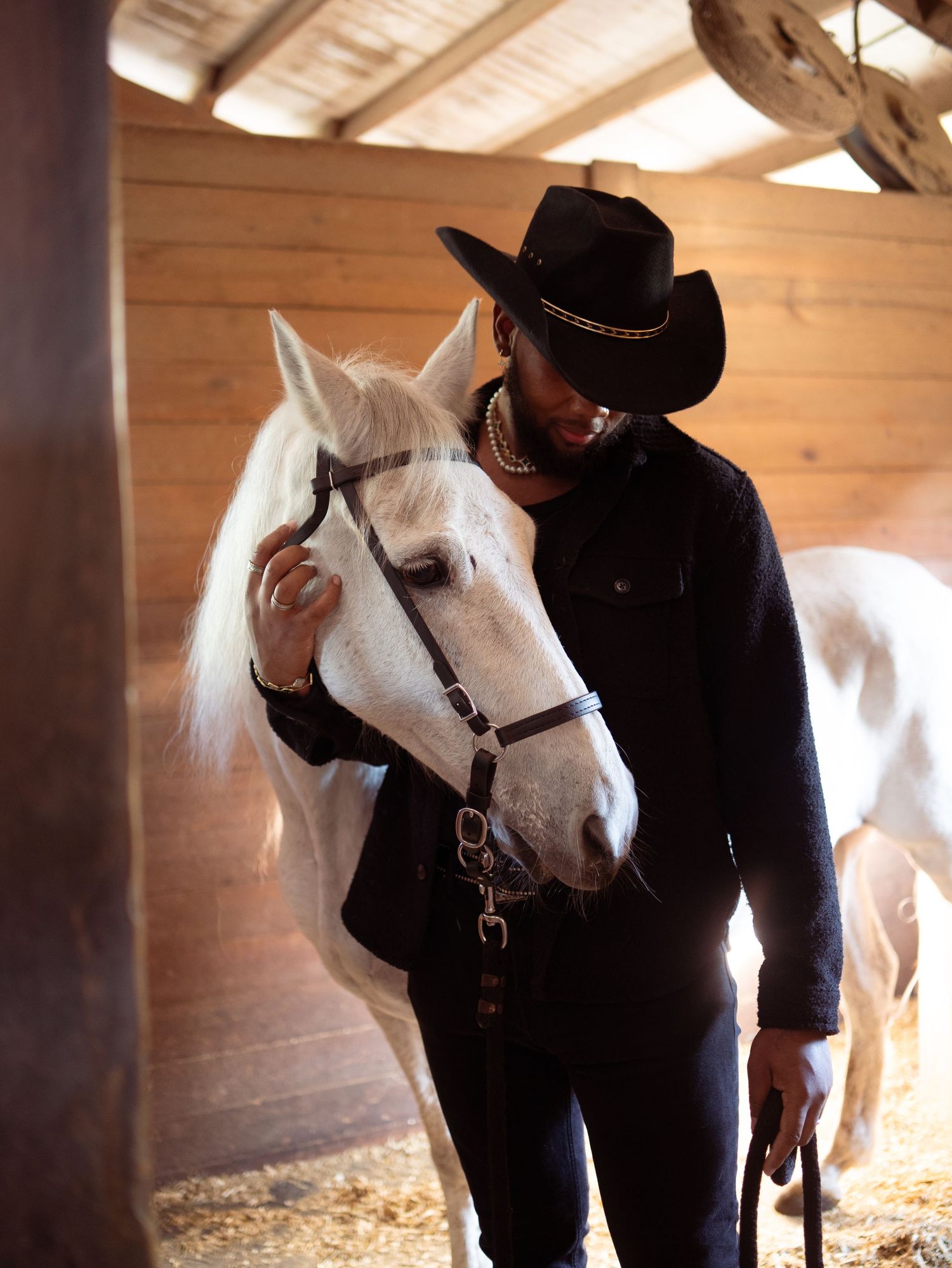 Person in black cowboy hat and outfit tenderly interacting with a white horse inside a wooden stable.