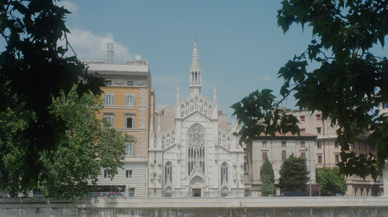 Gothic church facade with ornate spire framed by green trees under a bright blue sky in an urban European setting.