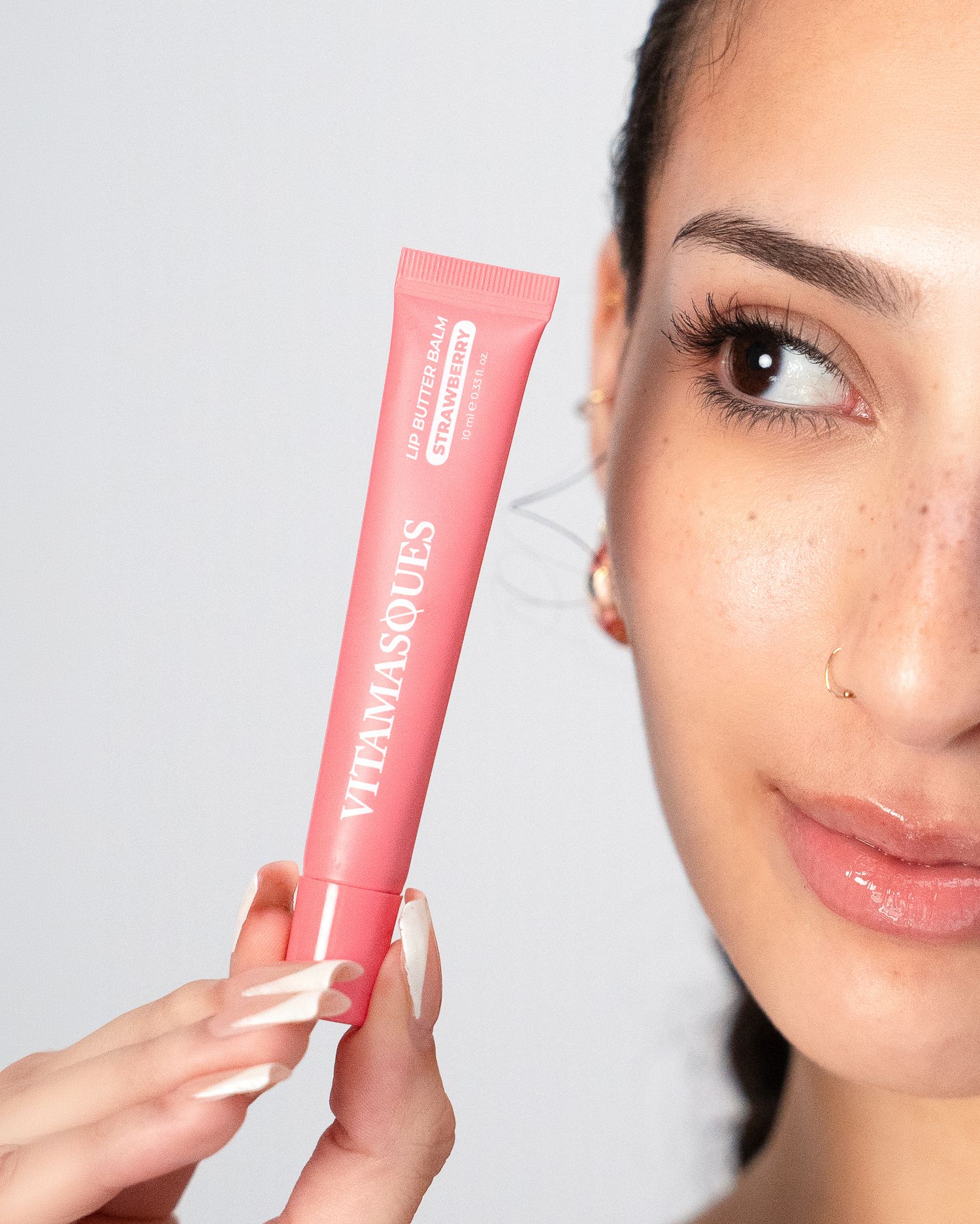 Woman holding a pink Vitamasques skincare tube next to her face against a light gray background.