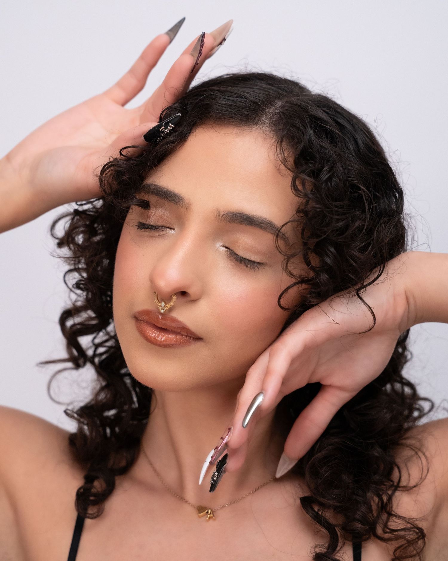 Young woman with curly hair and long nails poses gracefully with hands framing her face, wearing a gold nose ring.