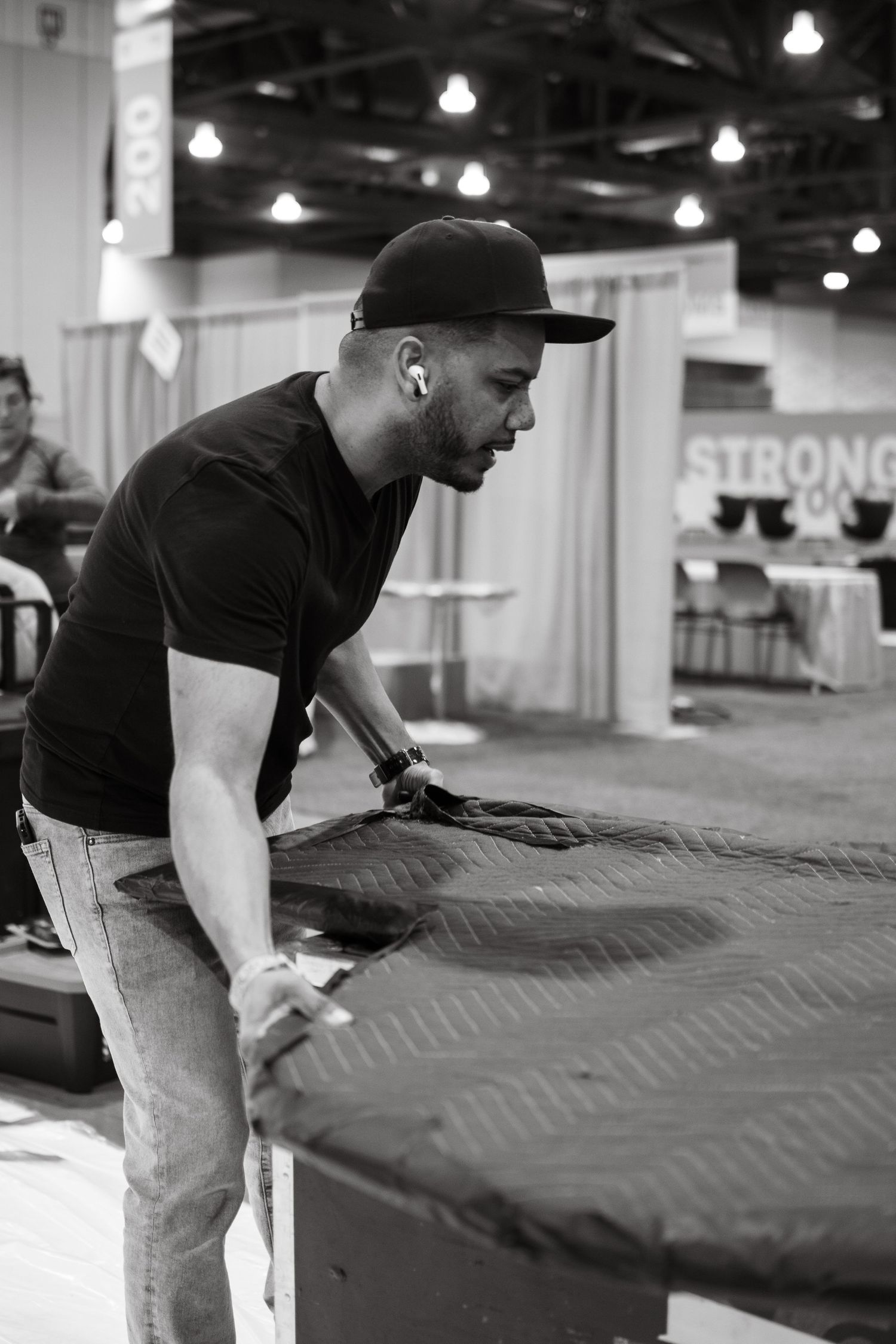 Man in black cap and t-shirt leaning over a padded surface at an indoor expo event, black and white photo.