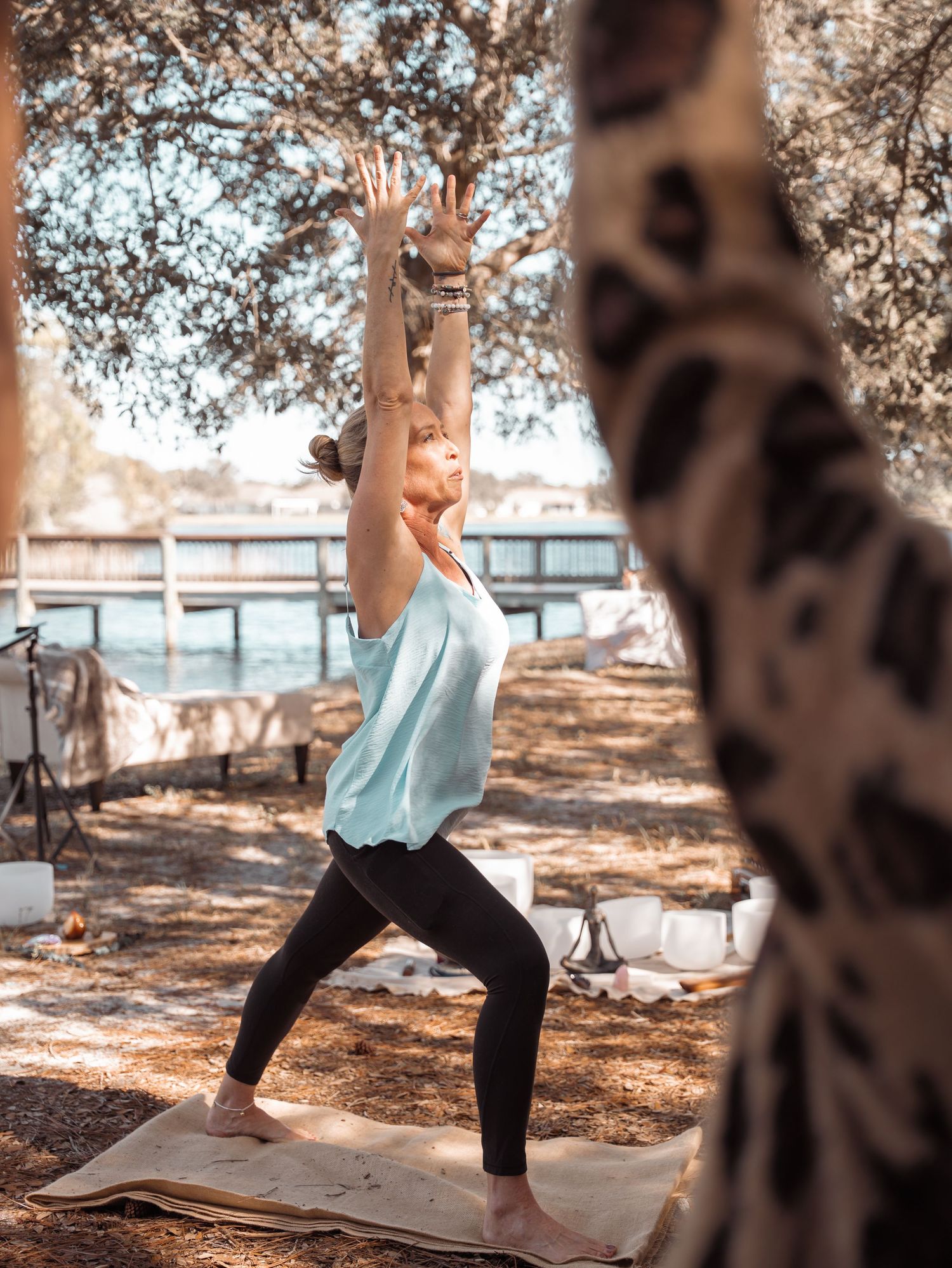 Woman practicing warrior yoga pose outdoors on a mat under trees near water, wearing teal top and black leggings.
