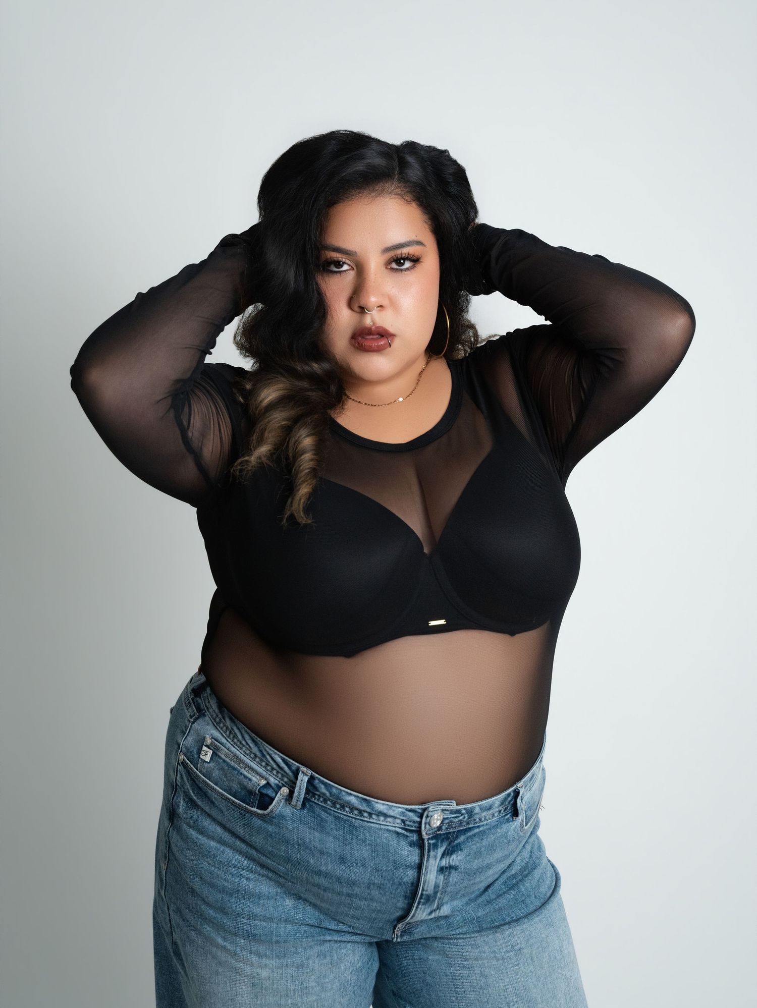 Plus-size model wearing black mesh long-sleeve top with structured bra and blue jeans against white background.