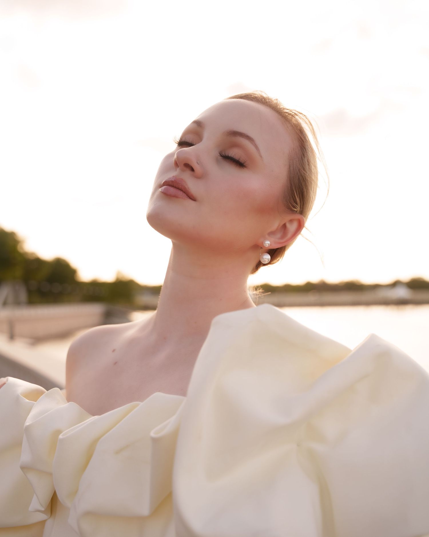 A woman in an off-shoulder cream gown poses gracefully outdoors at golden hour near a waterfront.
