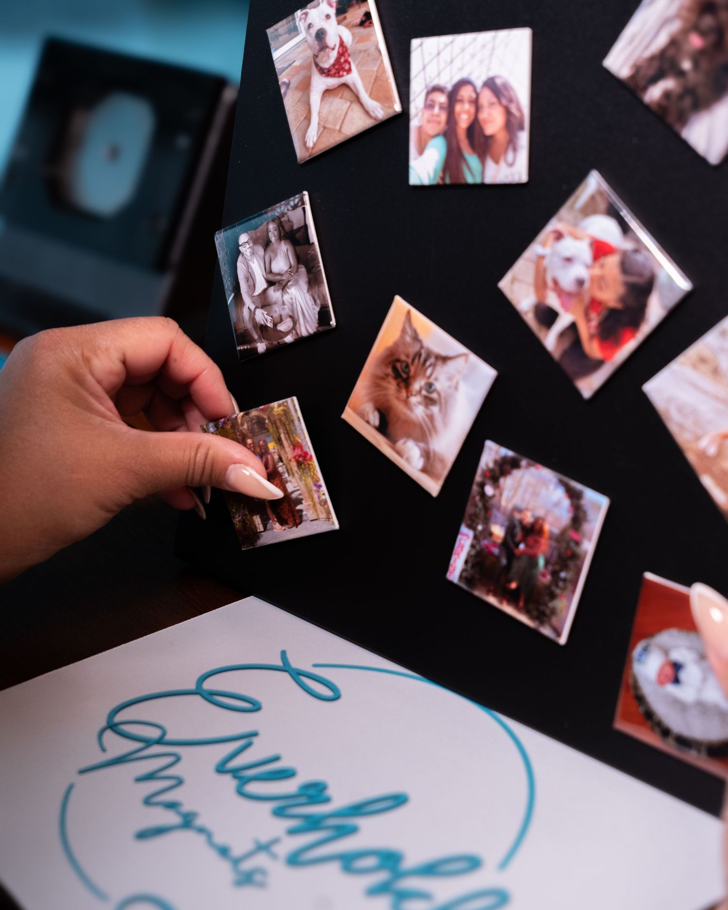 Hand placing small photo magnets on a dark board with colorful snapshots and Everhold cursive logo card nearby.