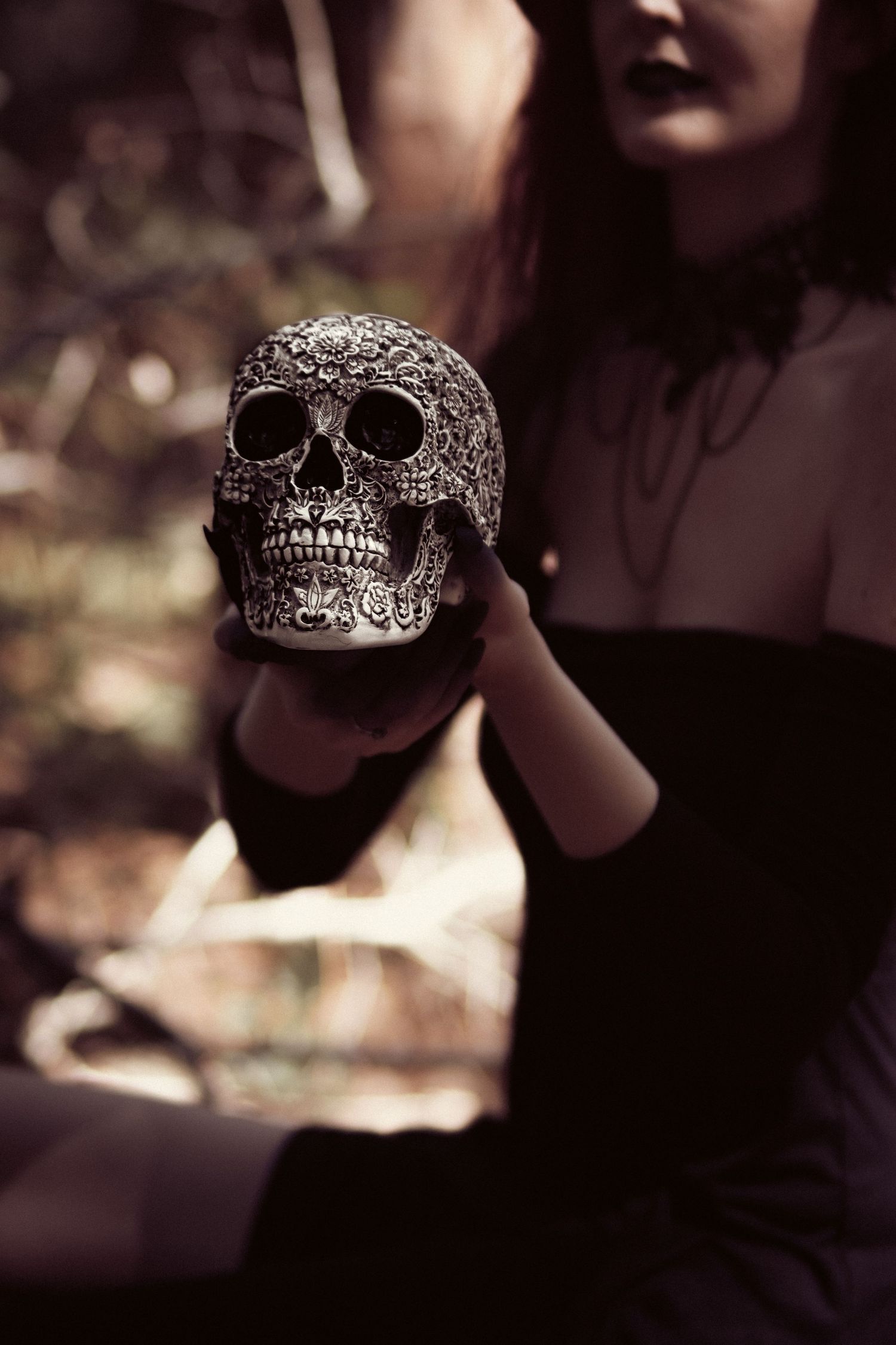 A gothic woman in black dress holds an ornate decorative skull in a dark, moody outdoor setting.