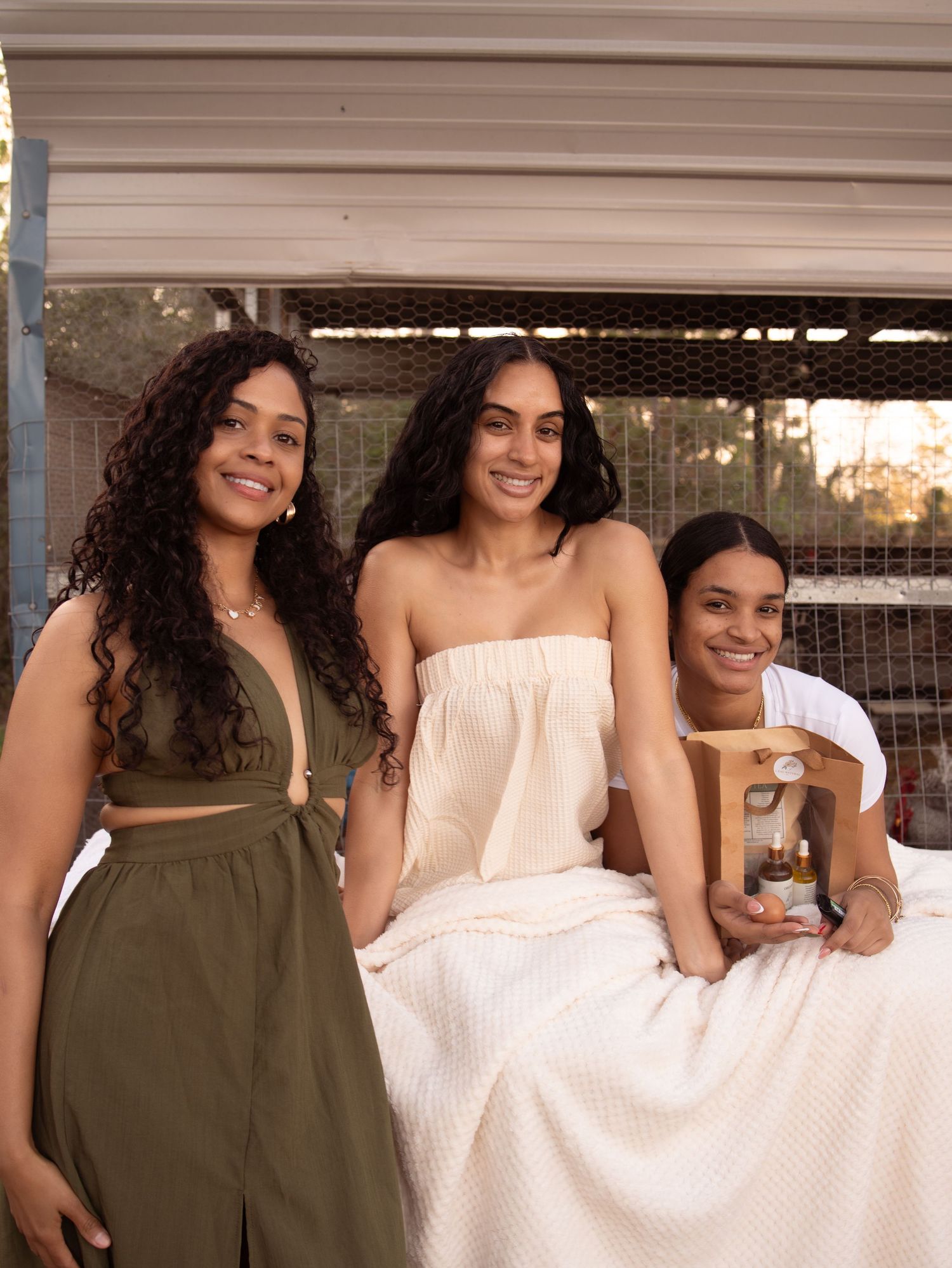 Three young women smiling together, one holding a beauty product gift set, wearing casual summer outfits outdoors.