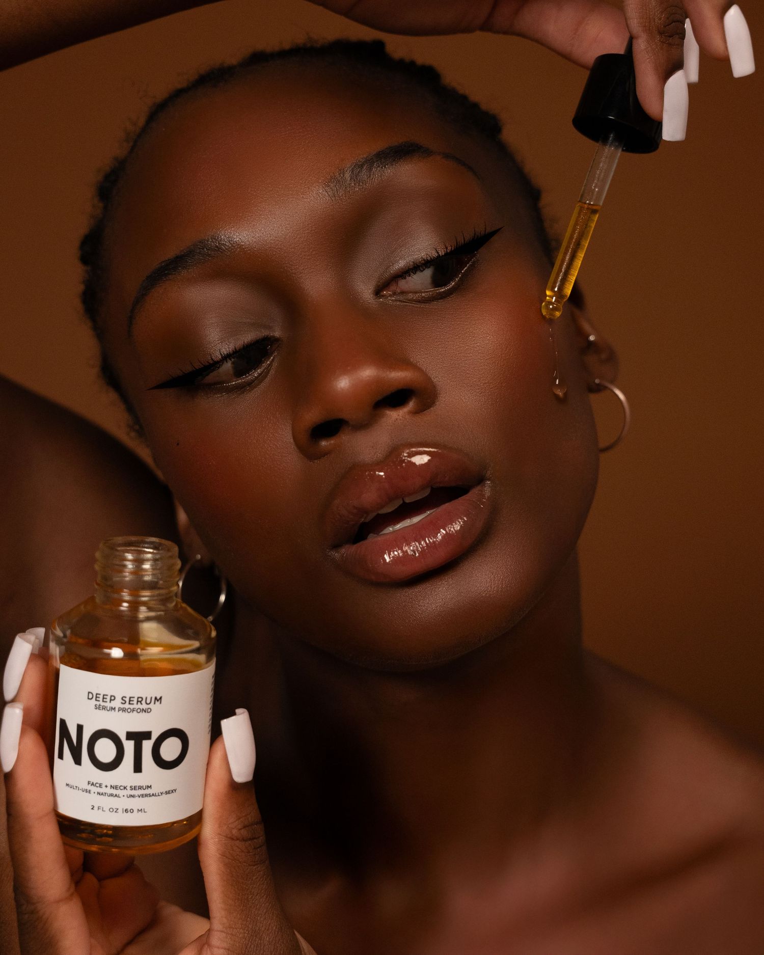 Model applying NOTO Deep Serum facial oil with dropper, showcasing glowing dark skin and bold eyeliner makeup look.