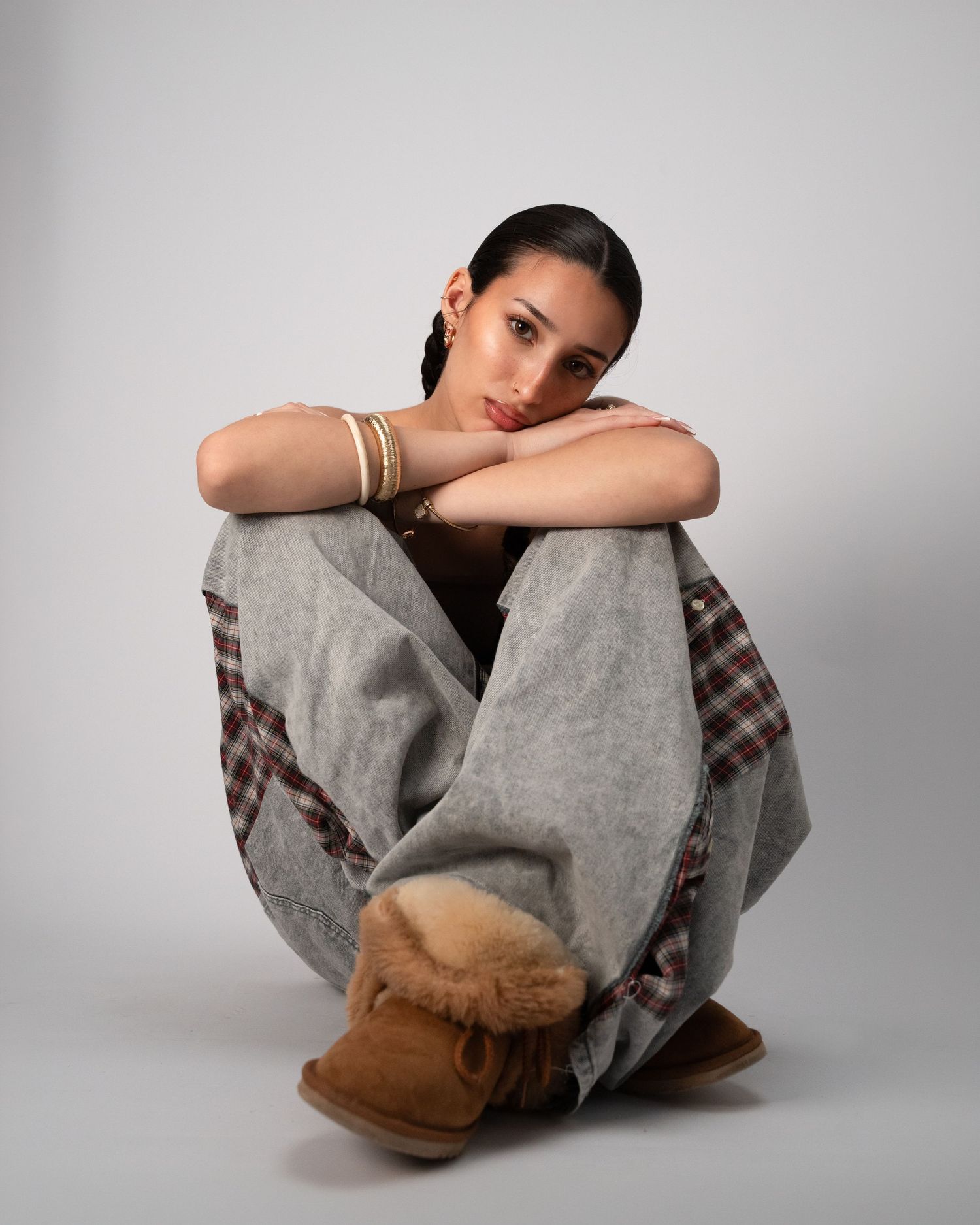 Young woman sitting on floor wearing gray wide-leg pants, plaid accents, UGG boots, and gold bracelets.