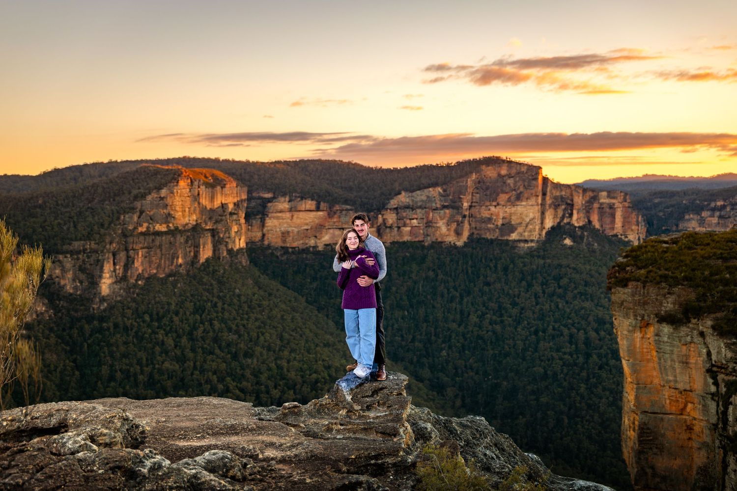 Couple standing at Walls Lookout at sunset overlooking the Blue Mountains valley with golden sky and sandstone cliffs.
