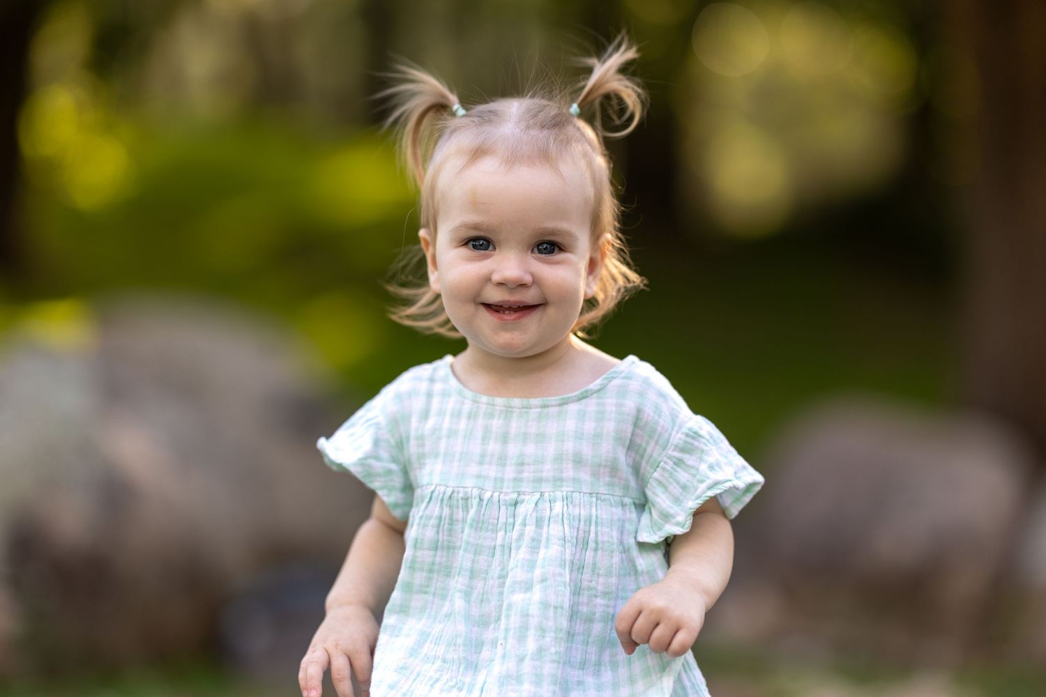 A young child in a light blue gingham dress poses playfully against a tree trunk in an outdoor setting at Tench Reserve, Nepean River.