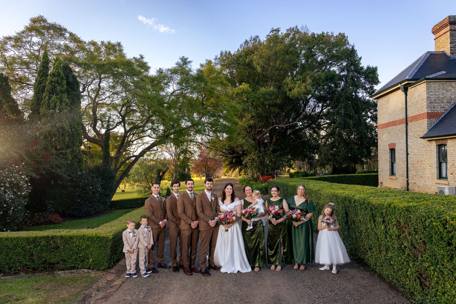 Wedding party poses outside Glenleigh Estate with lush hedges, groom in brown suit, bride in white, bridesmaids in green.