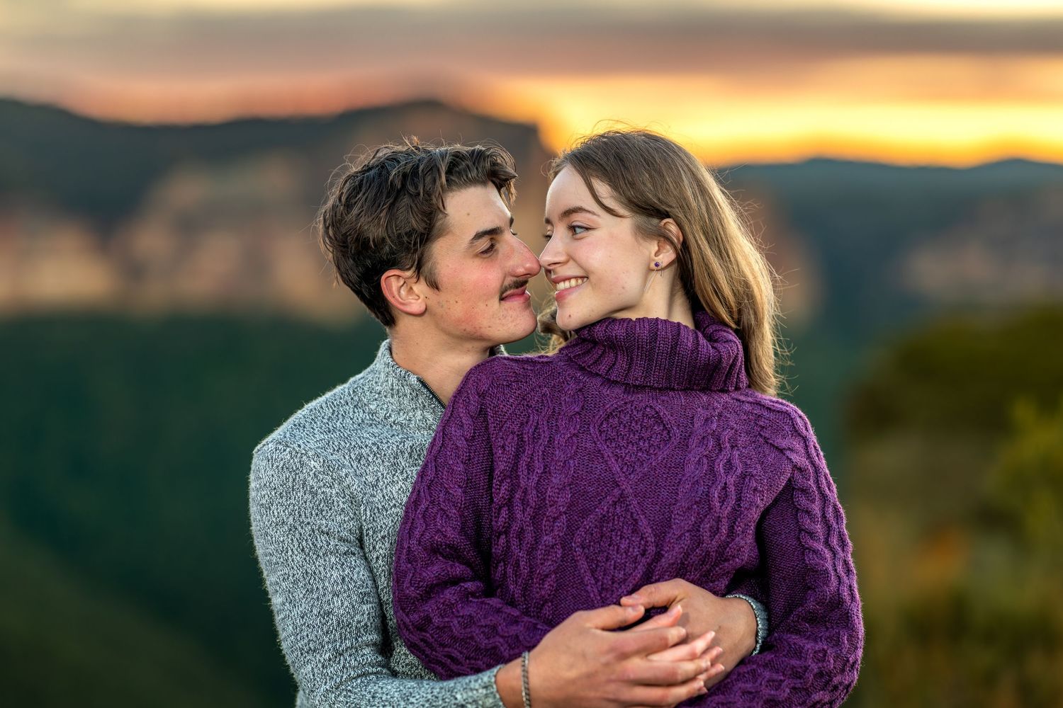 Couple embracing and smiling at sunset at Walls Lookout uwith mountain landscape in background, wearing cosy sweaters.