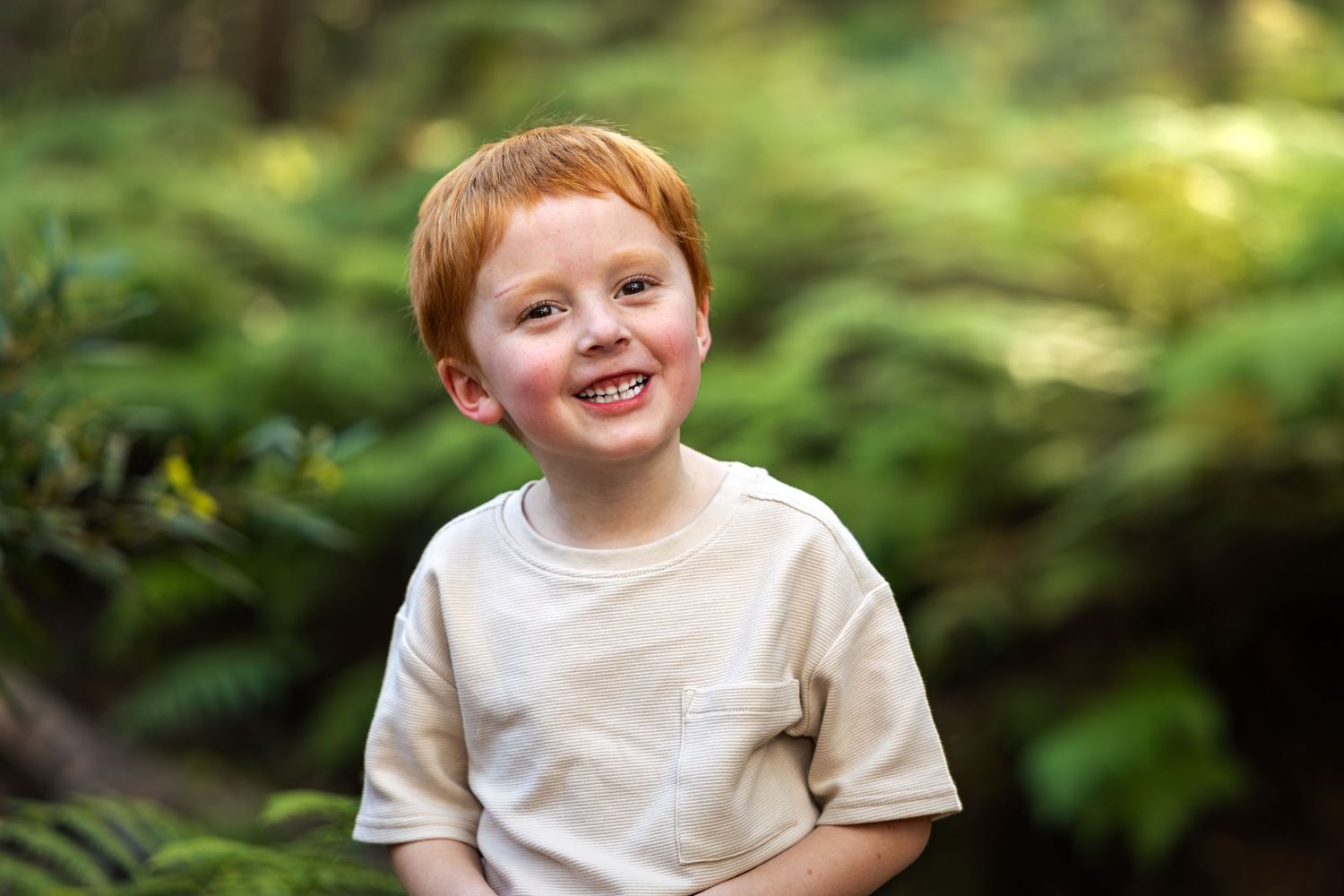 A cheerful red-haired boy smiling in a lush green forest, wearing a beige t-shirt.