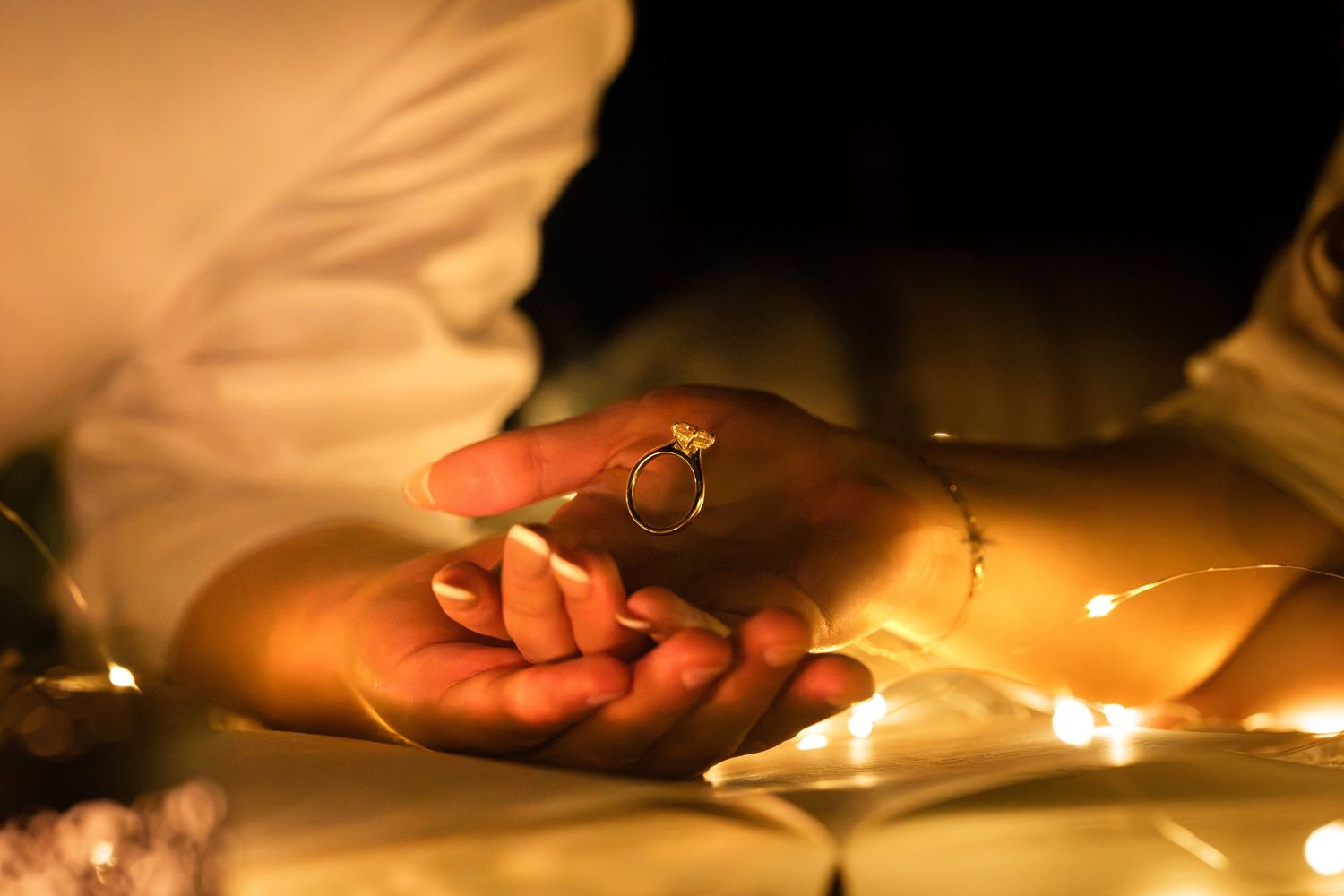 Hands holding a gold diamond ring surrounded by warm fairy lights in a romantic proposal setting.