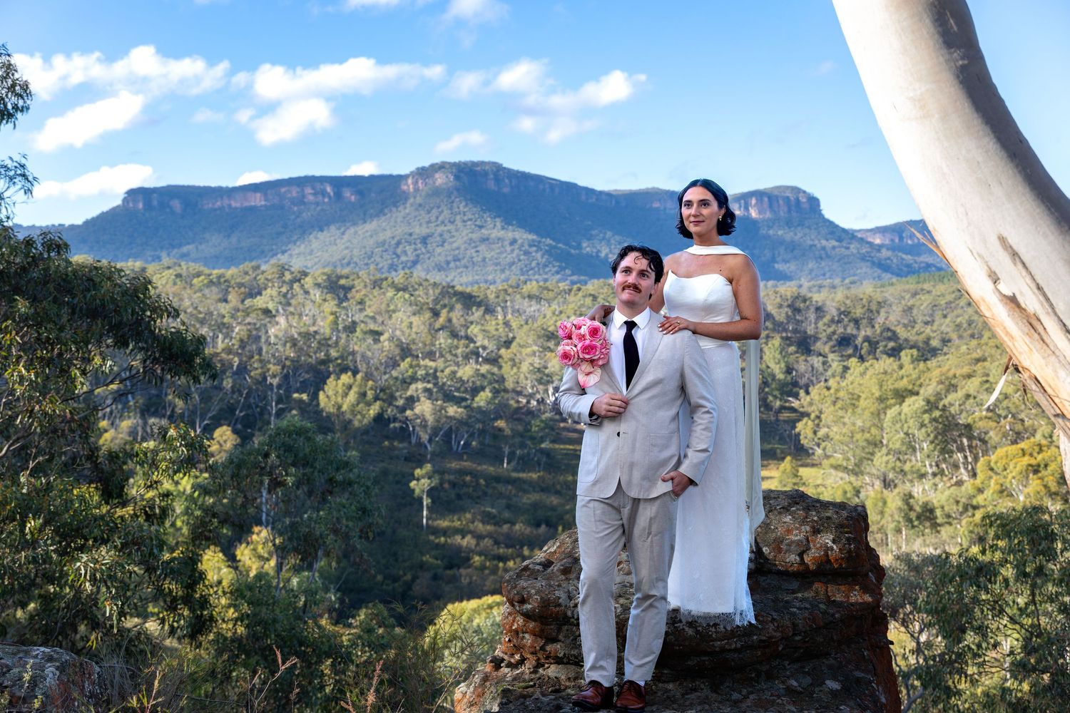 Couple in wedding attire posing on rocky outcrop with Blue Mountains landscape and dramatic cliffs in background at Maya Sanctuary.