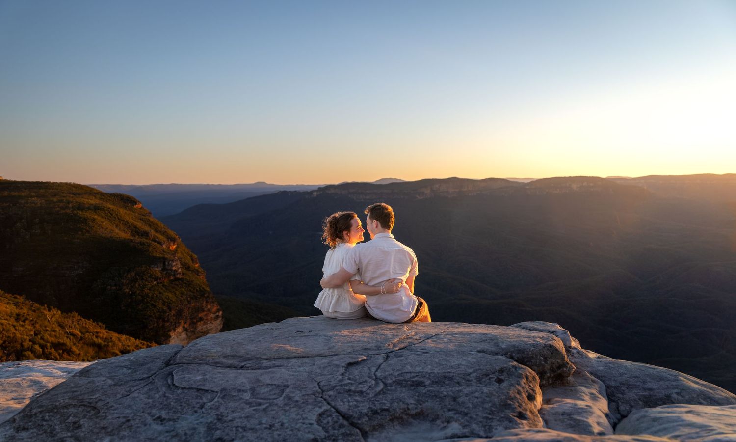 Couple sitting on rocky mountain overlook during golden sunset with panoramic views at Lincoln's Rock in Wentworth Falls, Blue Mountains.