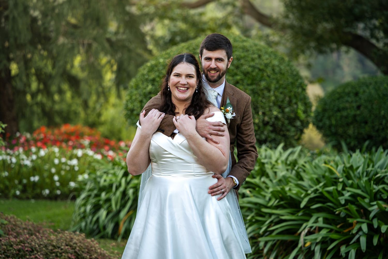 Bride and groom embrace in a lush garden, wearing wedding attire with colorful flowers in the background at Glenleigh Estate.