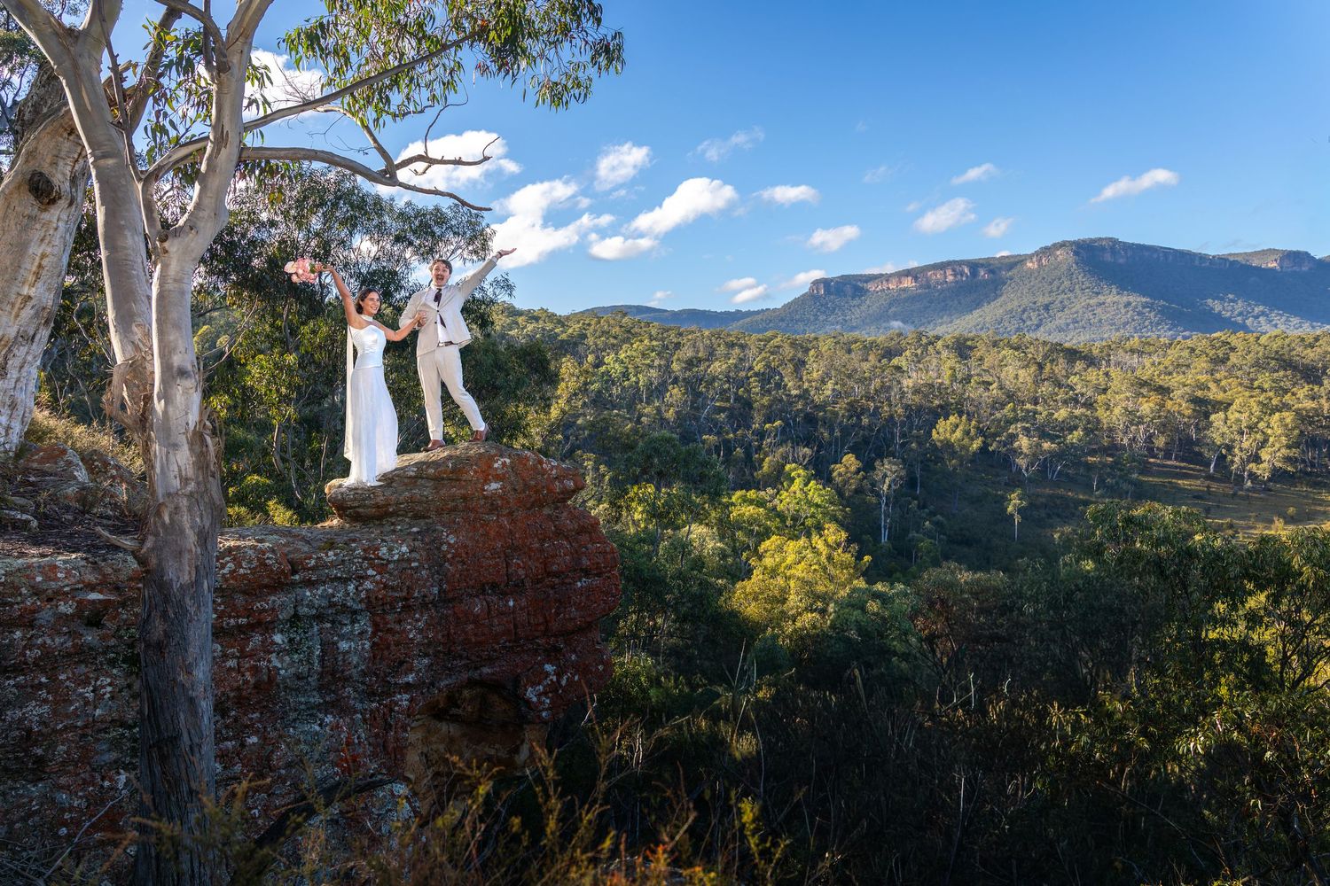 Couple in white wedding attire standing on a rocky cliff overlooking lush green valley and mountains at Maya Sanctuary.