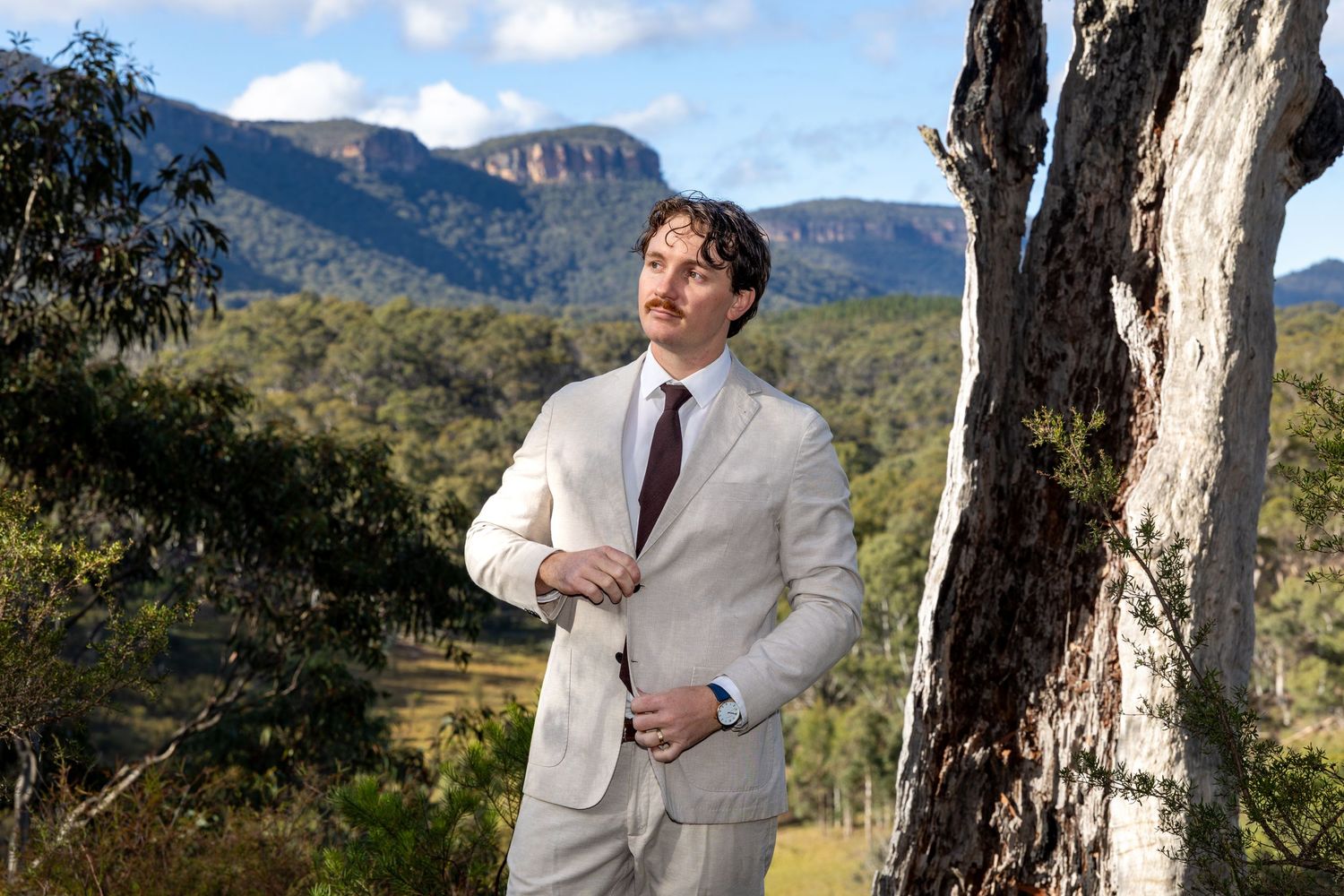 Man in cream suit standing by eucalyptus tree with Blue Mountains backdrop in Australia at Maya Sanctuary.