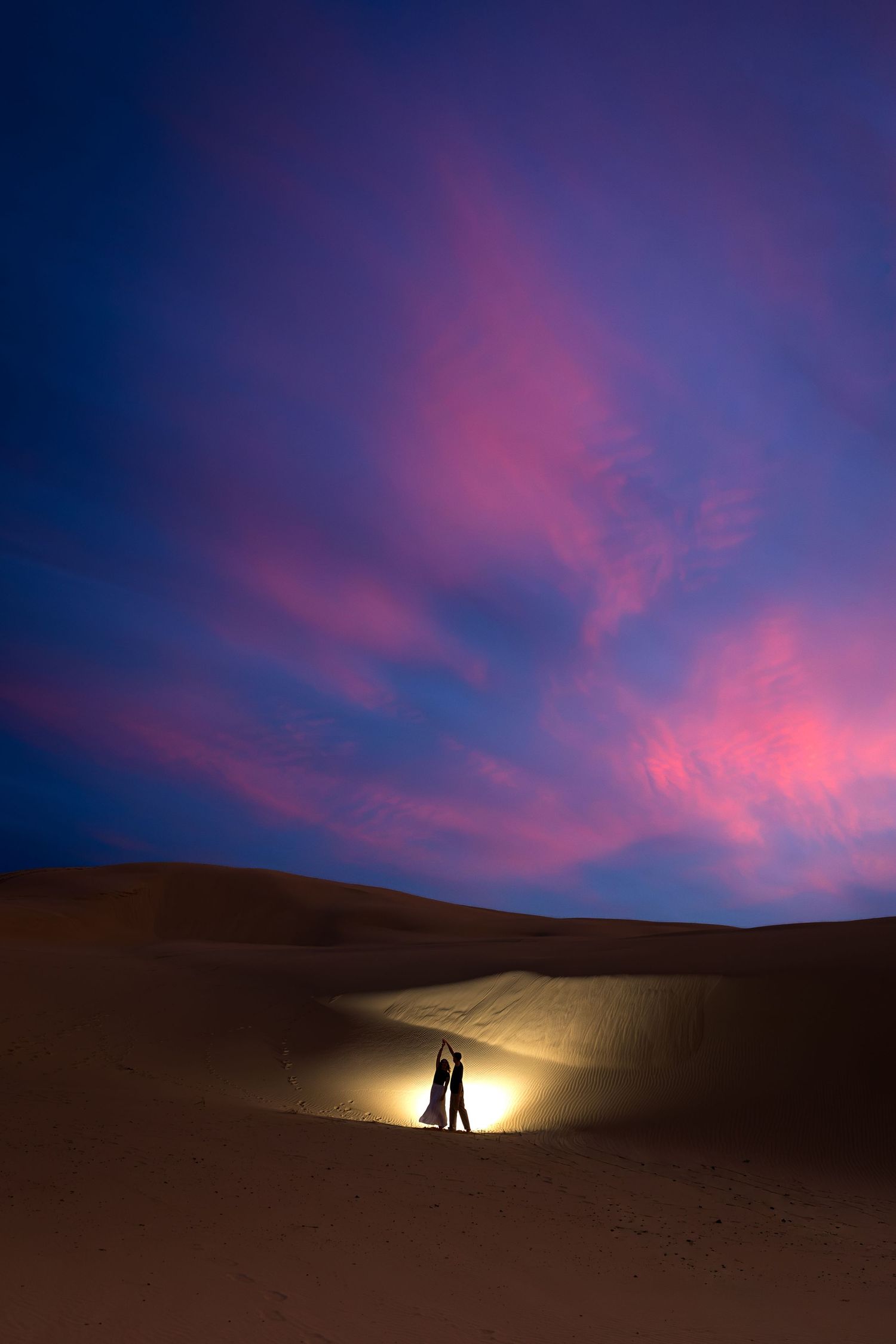 Couple silhouetted by glowing light on desert sand dunes under a dramatic pink and blue twilight sky.