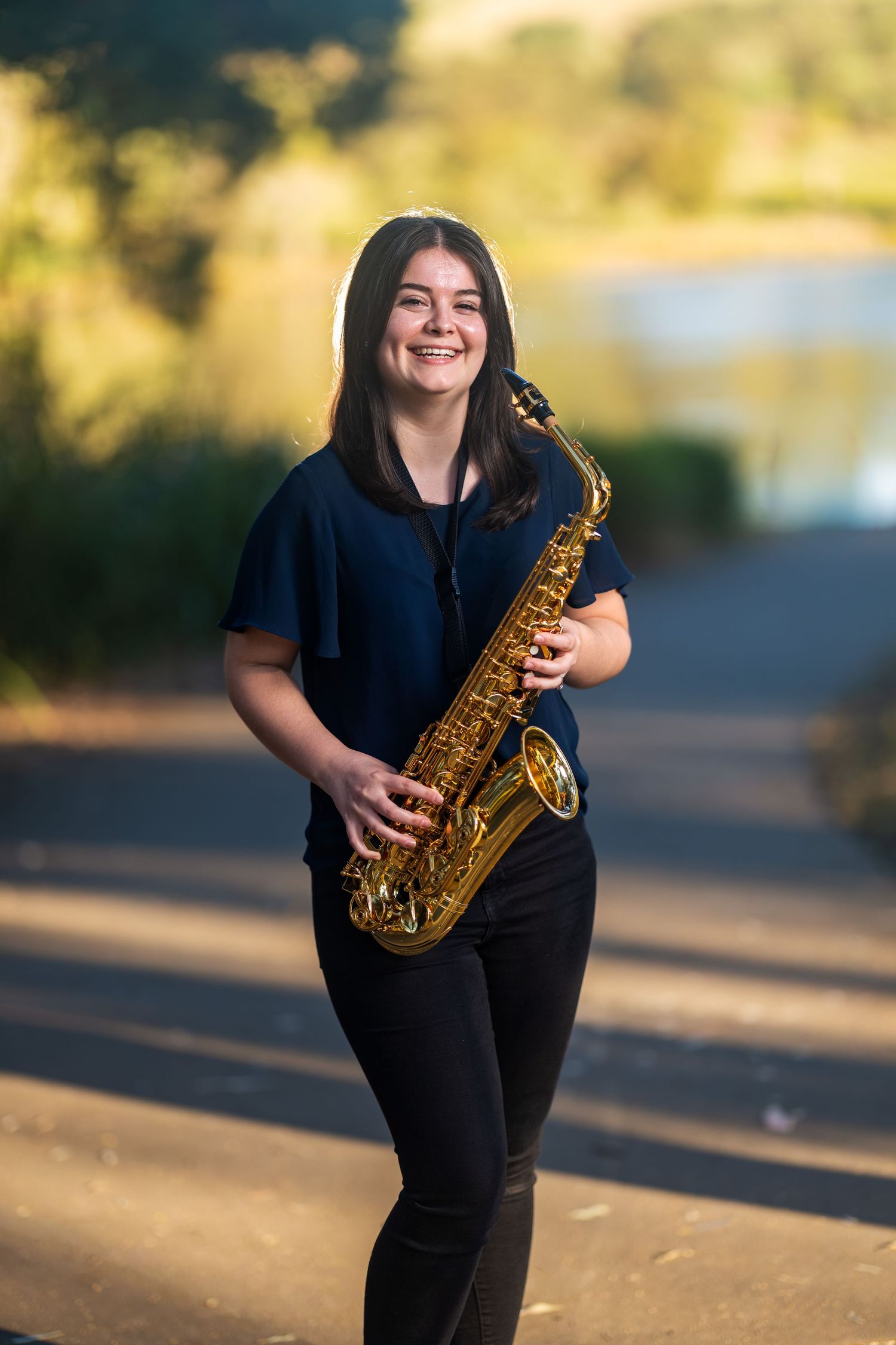 Smiling young woman holding a gold alto saxophone outdoors on a path with a lake and trees in the background.