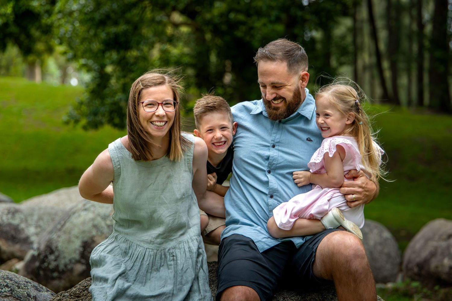 A happy family sits together outdoors on a rock, smiling and embracing in a natural green setting.