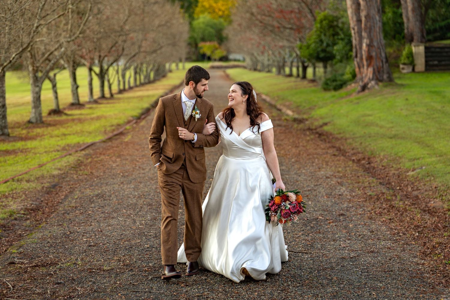 Bride and groom walking hand in hand down a tree-lined path at Glenleigh Estate, bride holding a colourful bouquet in a white gown.