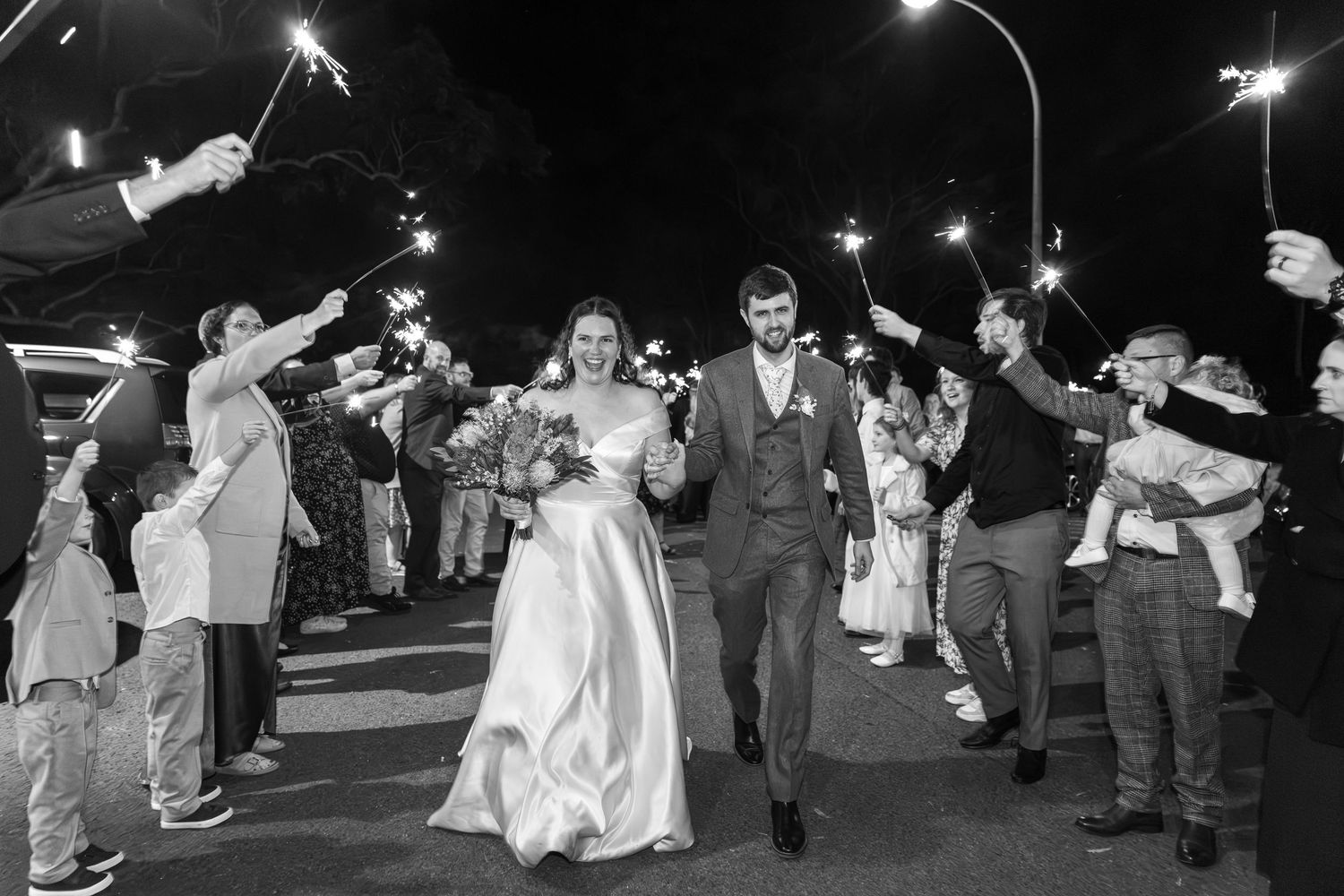 Black and white photo of newlyweds walking through sparkler send-off surrounded by cheering guests at night at Mulgoa Hall.